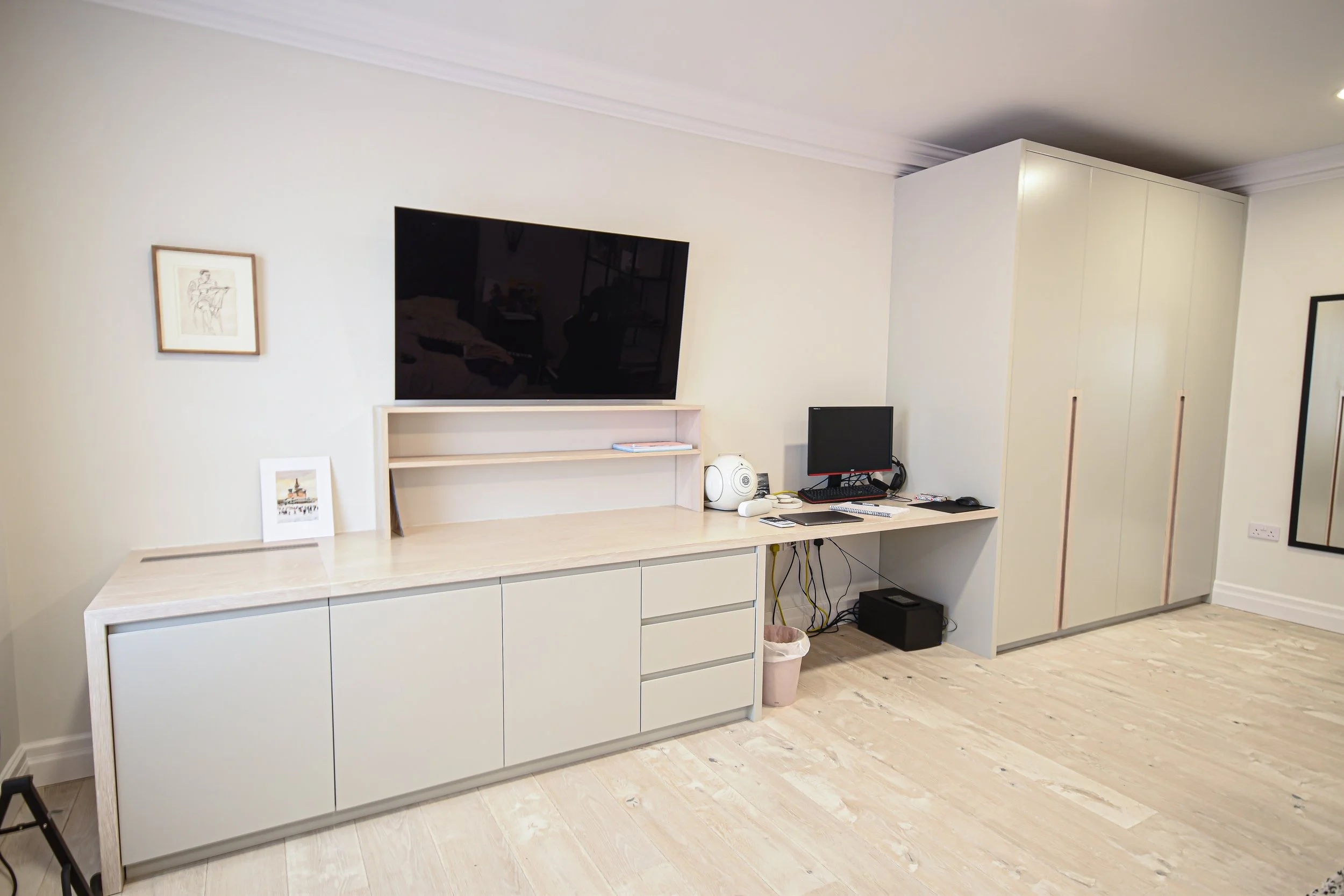 Minimalist home office with a TV, desk, and storage cabinets. Includes a computer monitor, speakers, framed art, and a wooden floor.