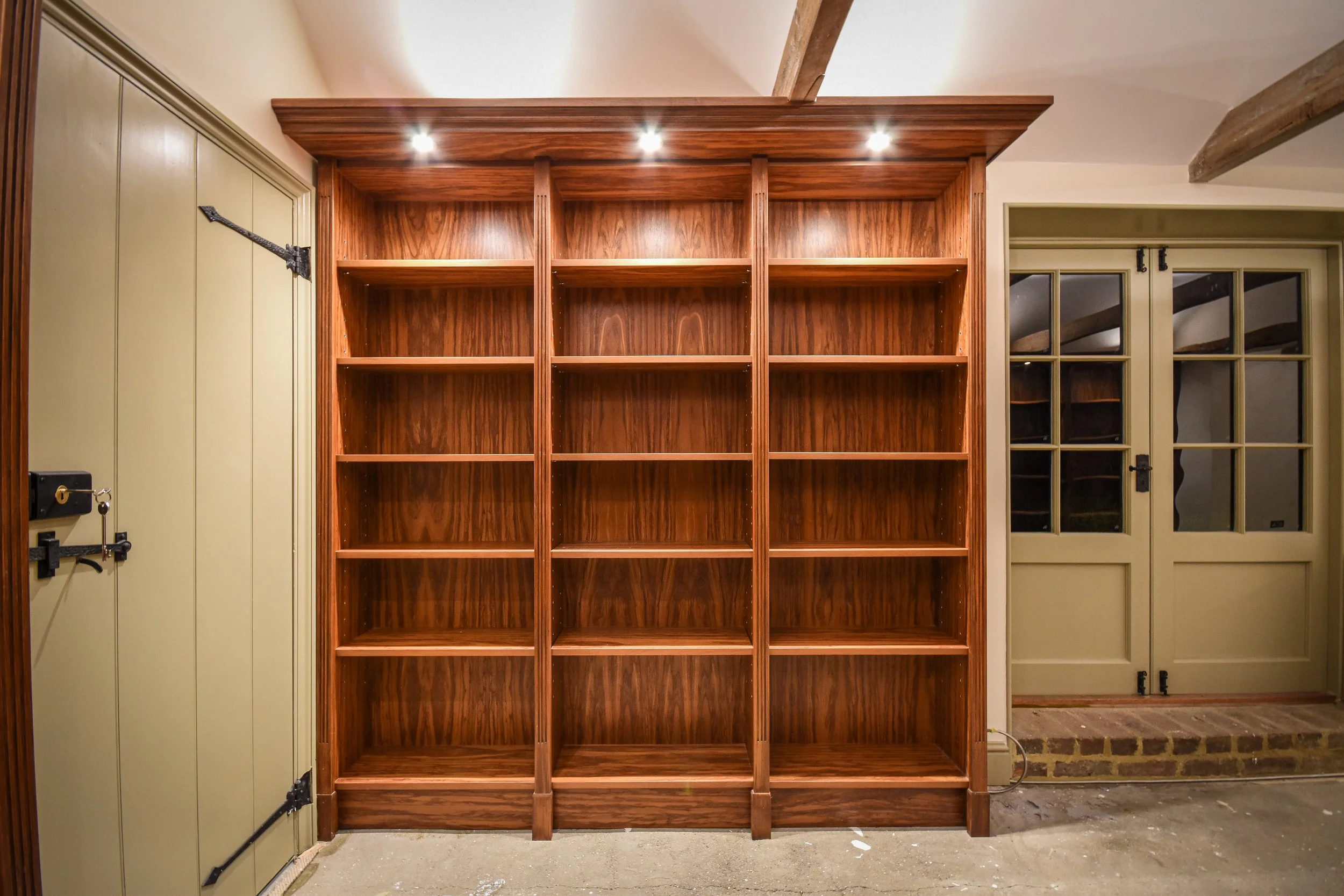 Built-in wooden bookshelf with empty shelves and overhead lighting, located between a wooden door and a pair of French doors in a room.