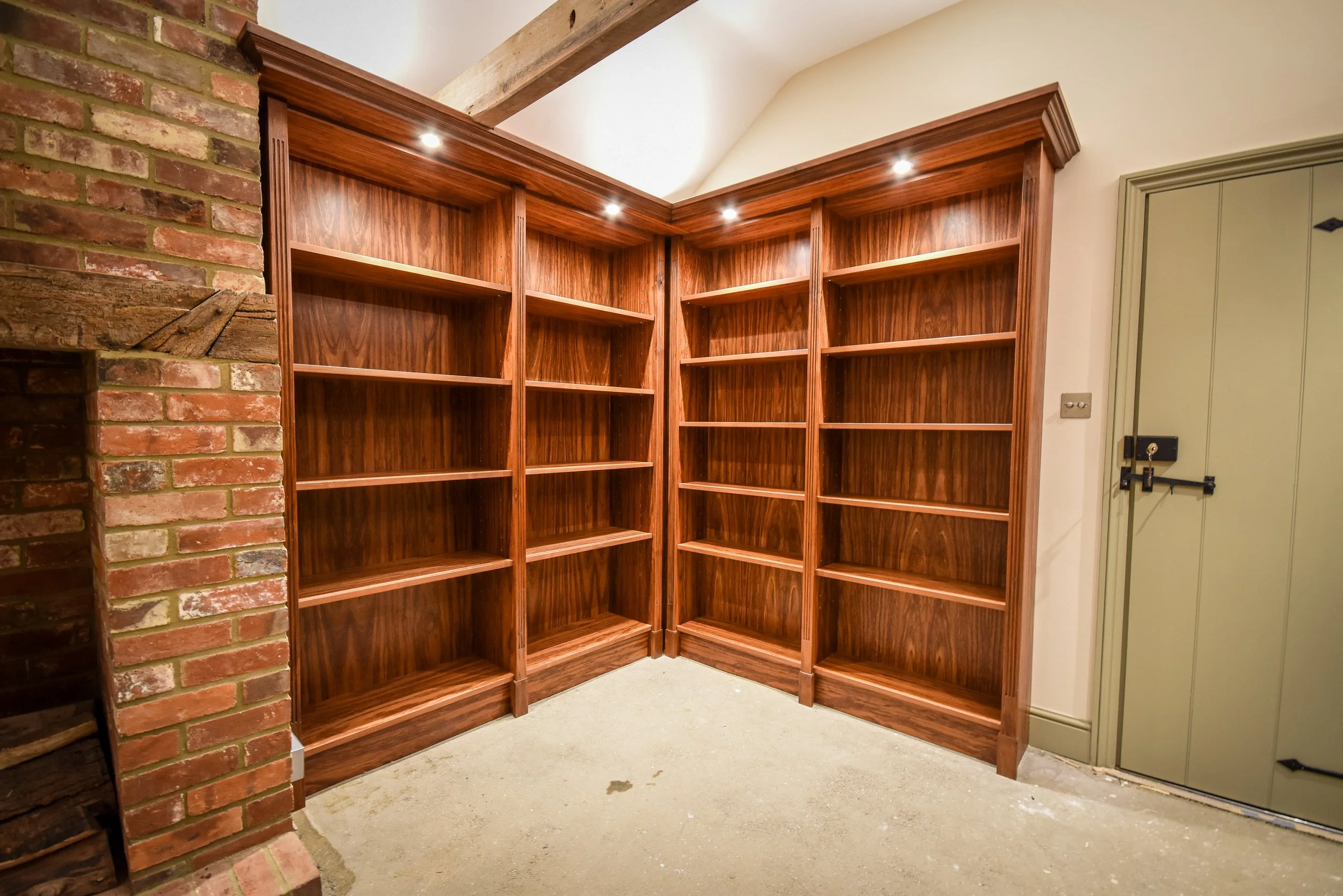 Empty wooden bookshelves in a corner with built-in spotlights, next to a brick fireplace and a closed wooden door.