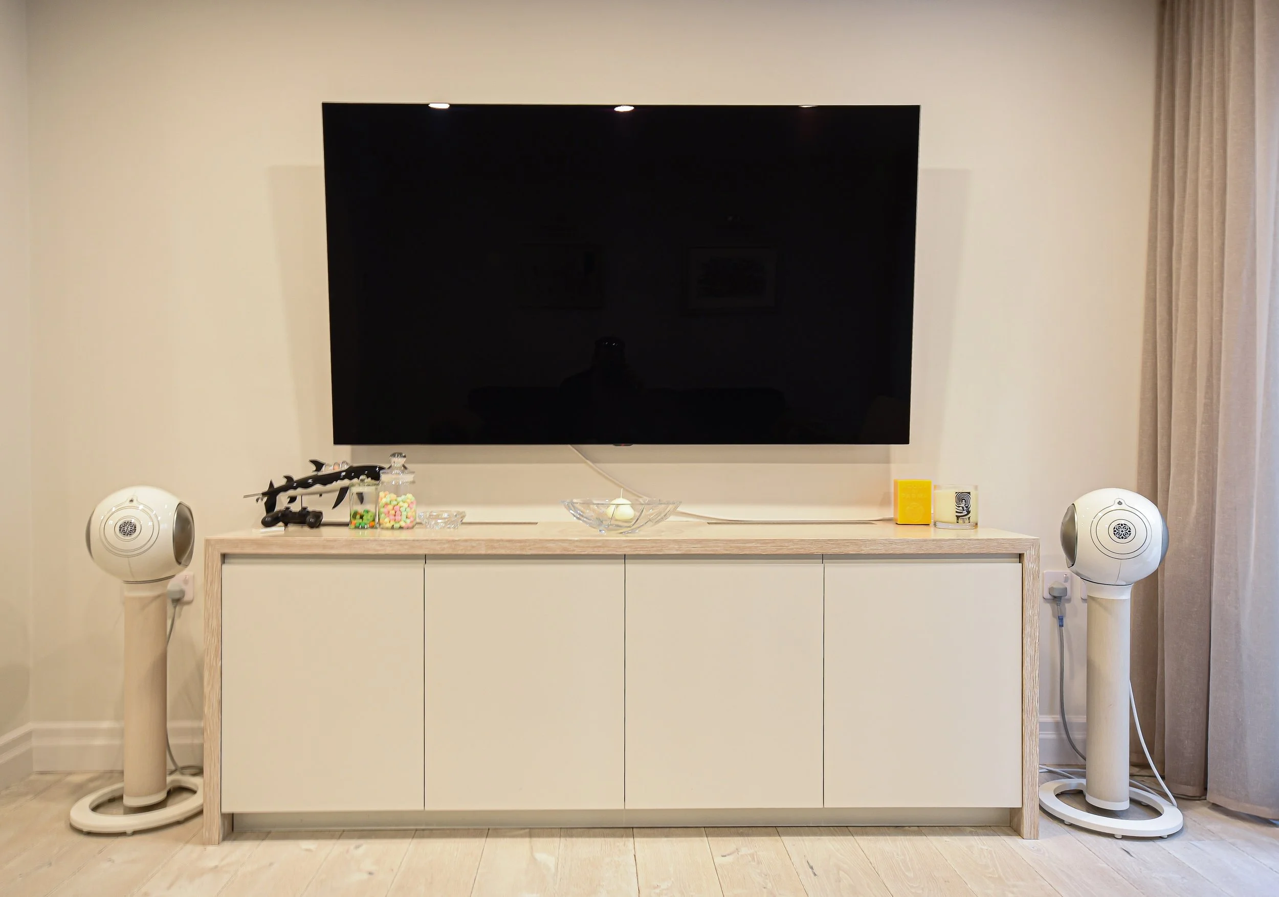 Living room setup with large TV mounted on wall, modern speakers, and wooden cabinet. Items on cabinet include decorative objects and containers.