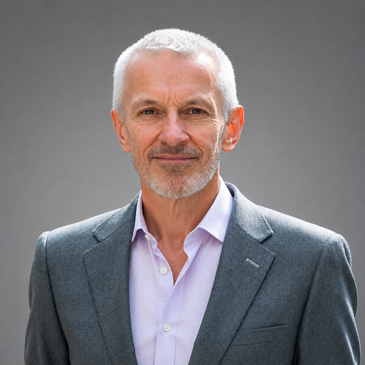 A mature man with gray hair and beard wearing a gray suit and light purple shirt, standing against a plain gray background.