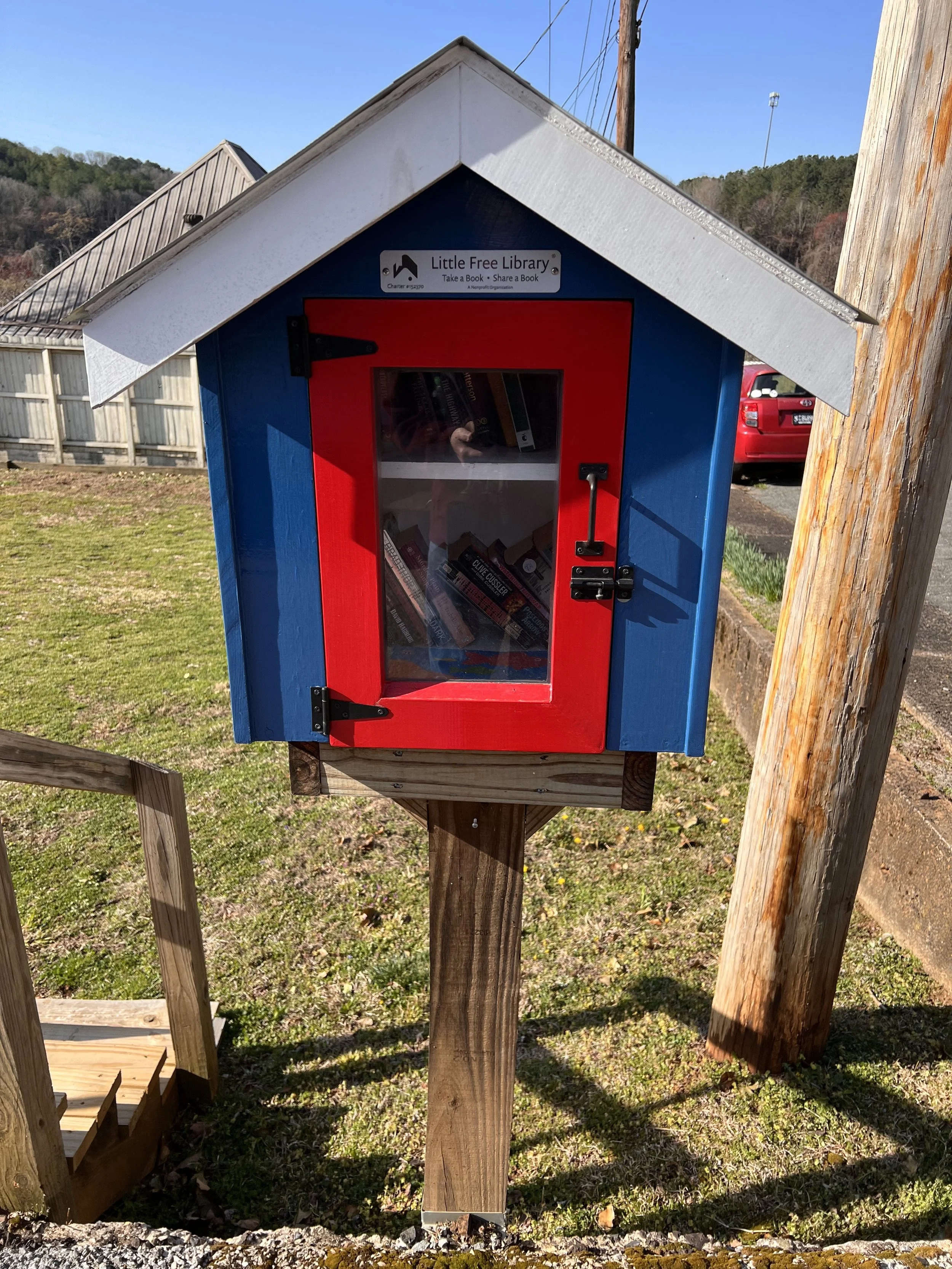 Little Free Library structure with a blue exterior and red door, filled with books, mounted on a wooden post, located in a grassy area near a wooden fence and red car.