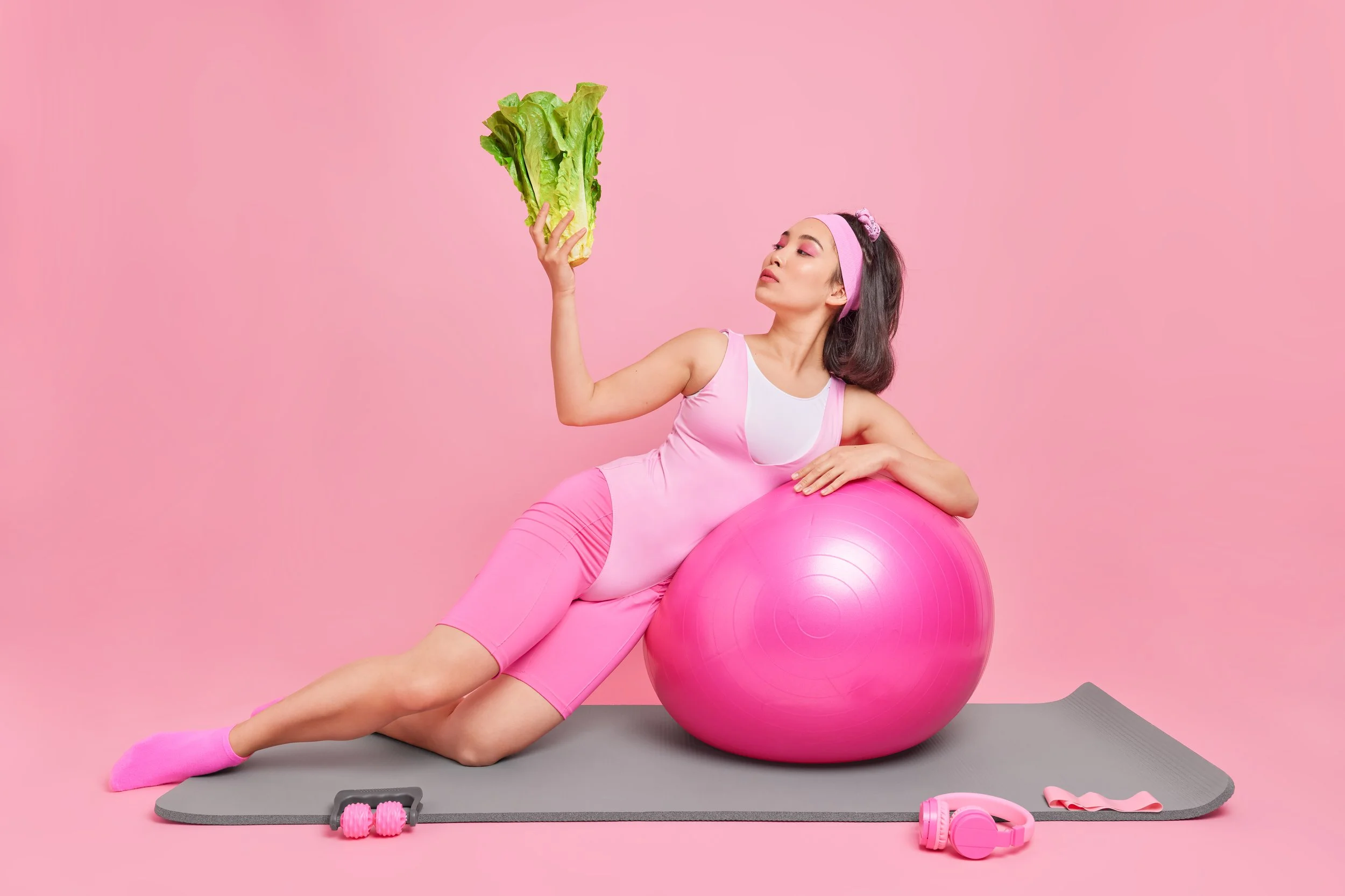 Woman in pink activewear holding lettuce while leaning on exercise ball, symbolising the connection between fitness and fibre-rich nutrition for recovery