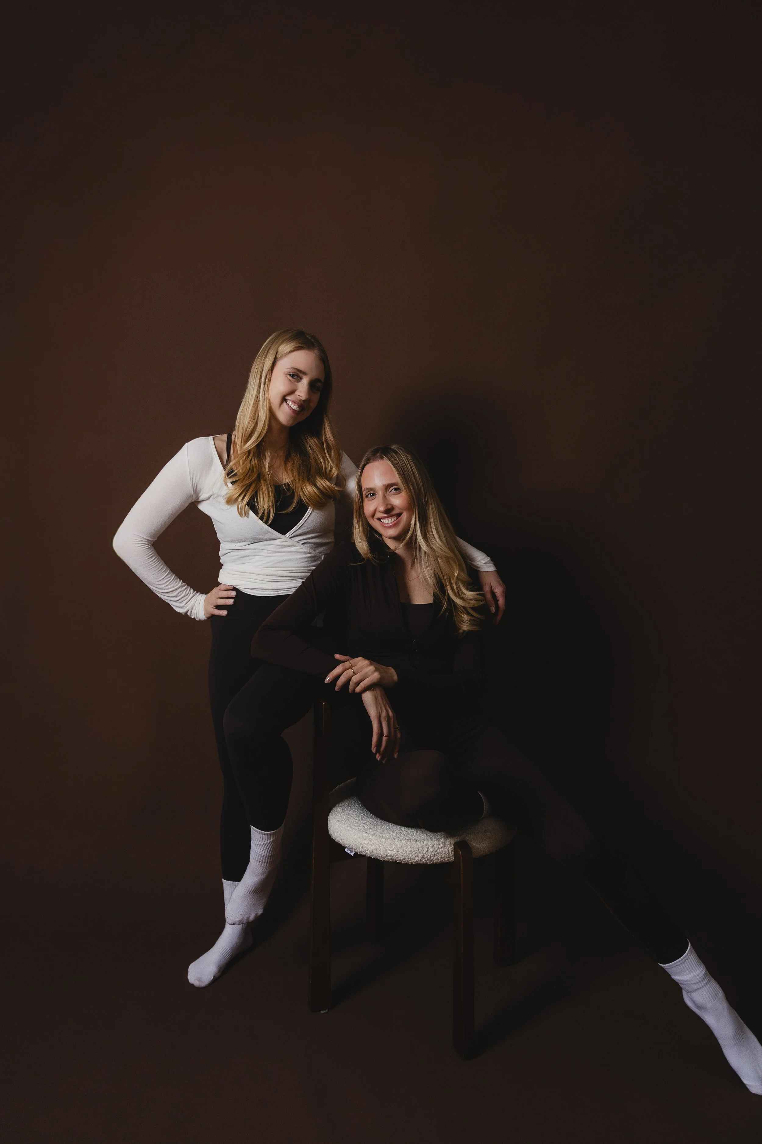 Zwei junge Frauen posieren vor einem dunklen Hintergrund. Eine steht links, trägt einen weißen Pullover mit schwarzem Top darunter, schwarze Hosen und weiße Socken. Die andere sitzt auf einem Stuhl, trägt einen schwarzen Anzug, schwarze Hose, weiße Socken und hat lange blonde Haare.
