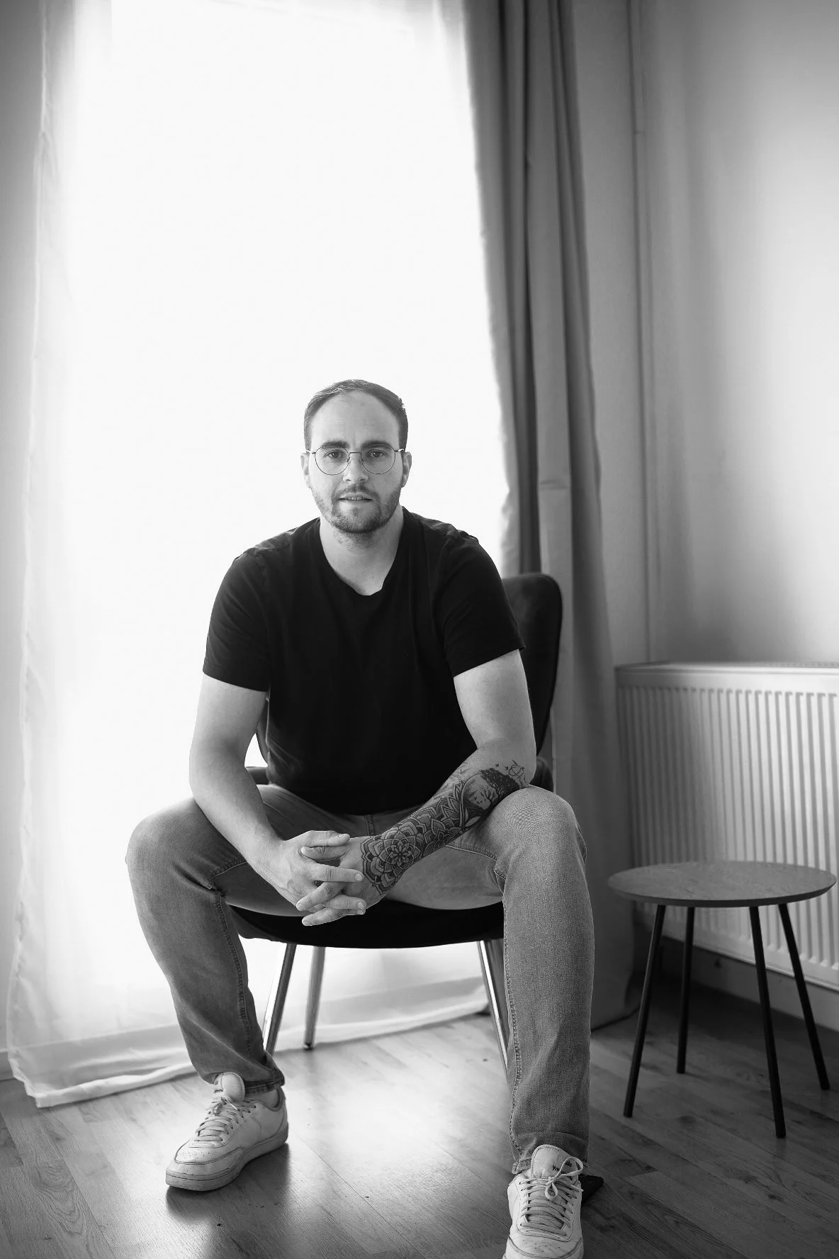 Schwarz-weißer Portrait eines Mannes, der auf einem Stuhl sitzt, trägt Brille und T-Shirt, hat ein Tattoo am linken Arm, befindet sich in einem Raum mit Vorhängen und Heizkörper.