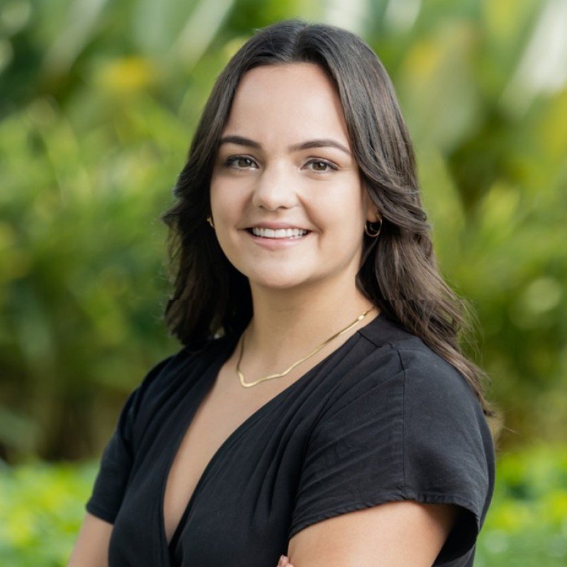 Young woman with dark hair smiling outdoors with green blurred foliage in the background.