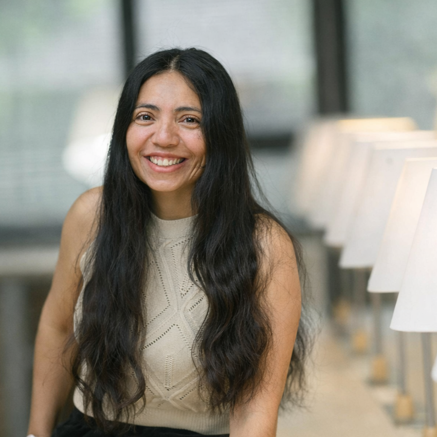 Une femme souriante avec de longs cheveux noirs, assise dans un lieu lumineux, entourée de lampes