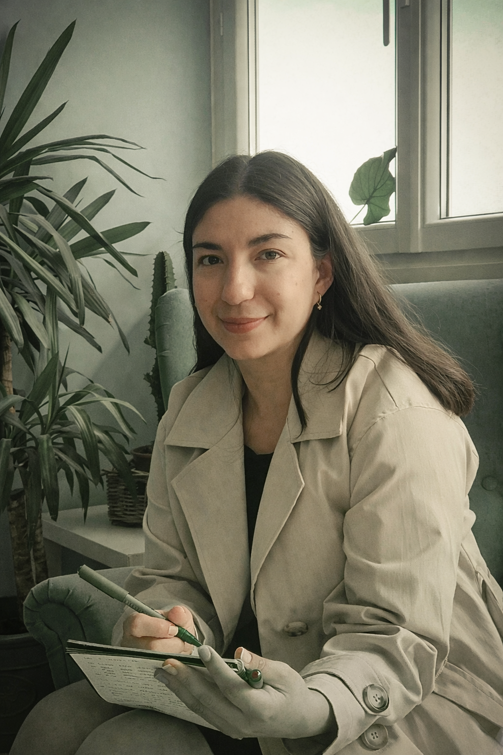 Jeune femme assise sur un fauteuil, écrivant sur un carnet, entourée de plantes vertes, dans une pièce lumineuse.