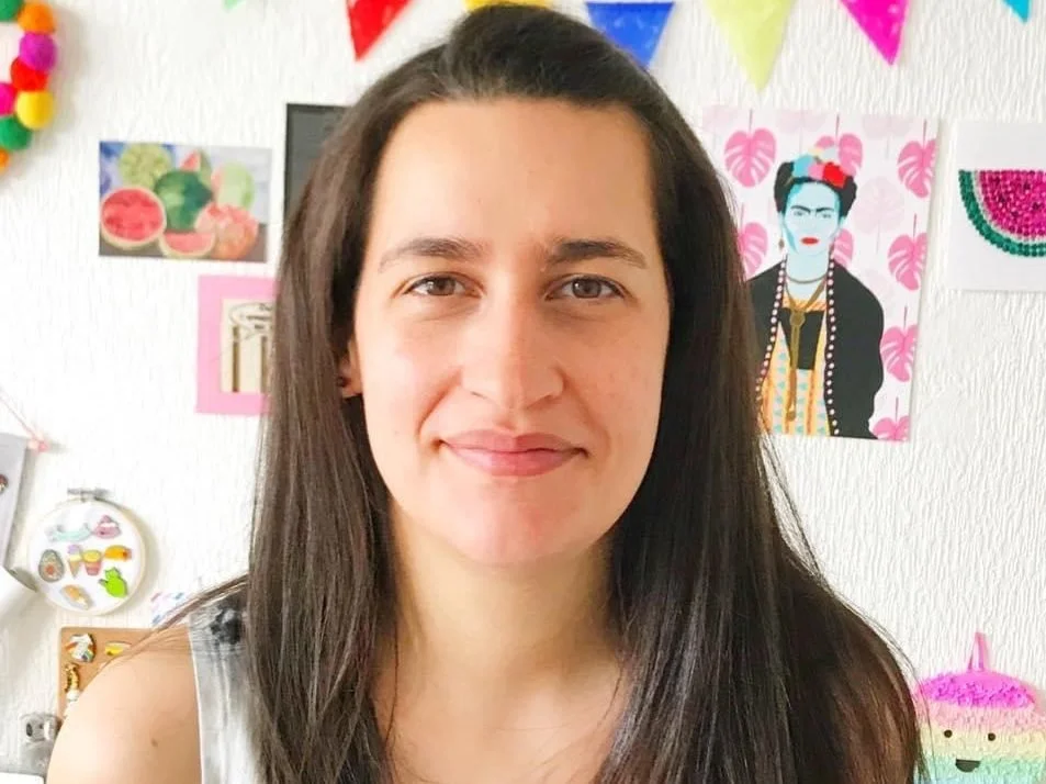 A person with long dark hair, smiling slightly, in front of a white wall decorated with colorful artwork, including an illustration of Frida Kahlo and abstract drawings. The room is bright and festive, with a cheerful ambiance.