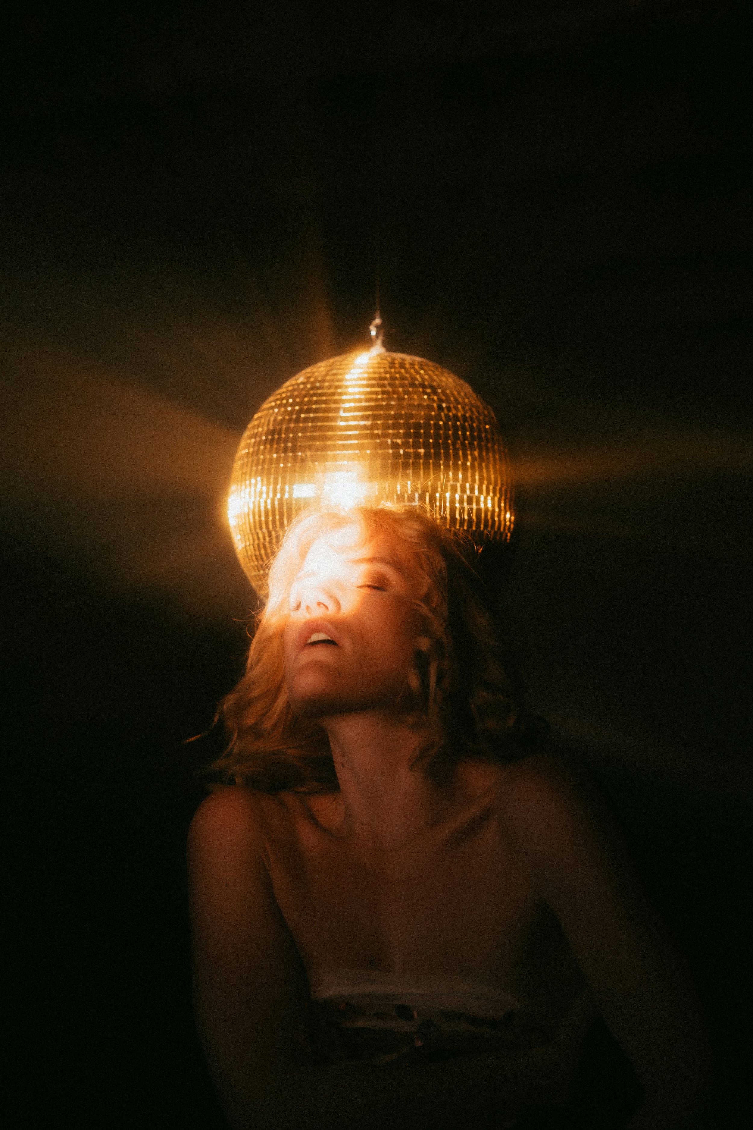 Une femme aux cheveux ondulés à la lumière d'un grand miroir disco doré suspendu au plafond, dans un endroit sombre.