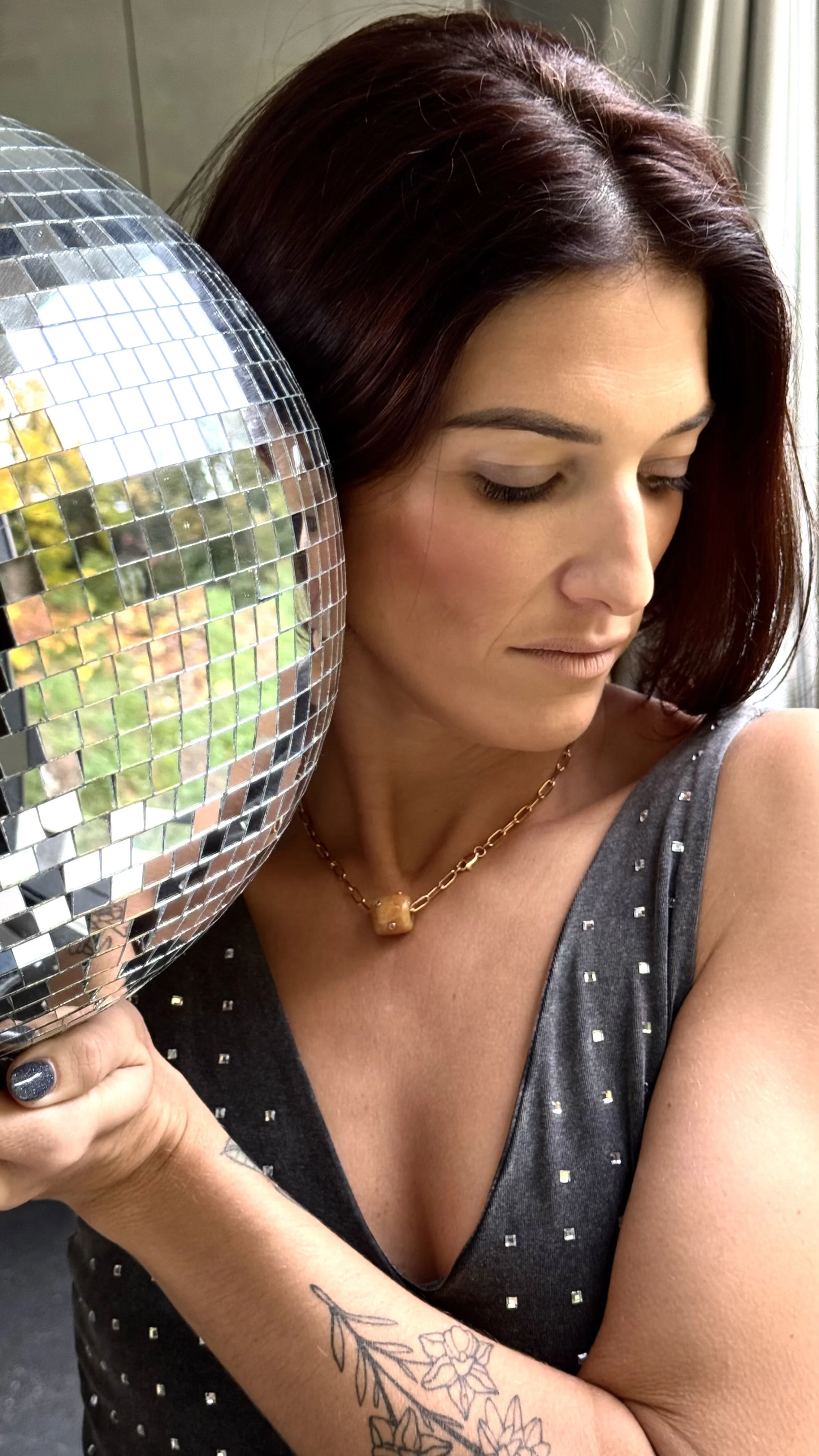 Femme avec tatouages et collier, tenant une boule disco, regardant vers le bas, en intérieur.