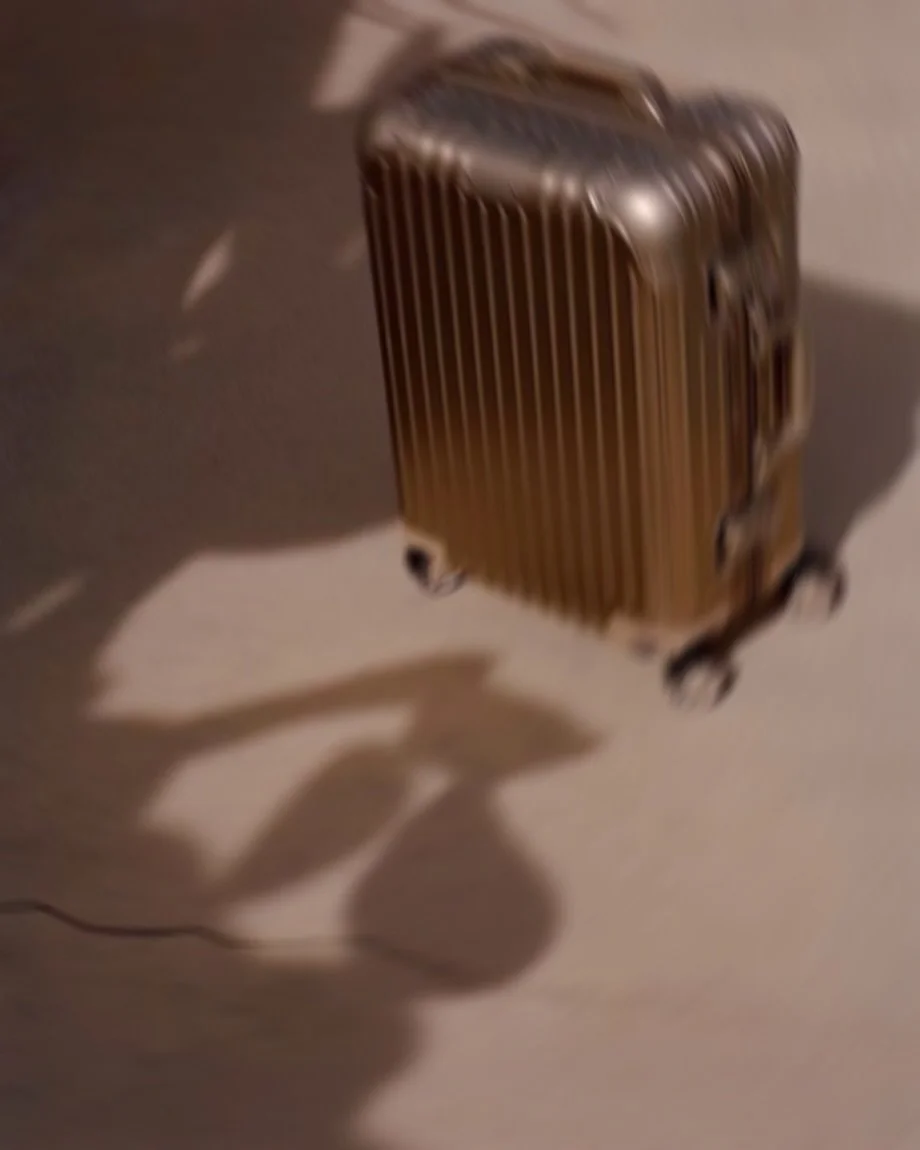 Blurred image of a metallic rolling suitcase in motion on a light-colored background, with a shadow cast on the ground.