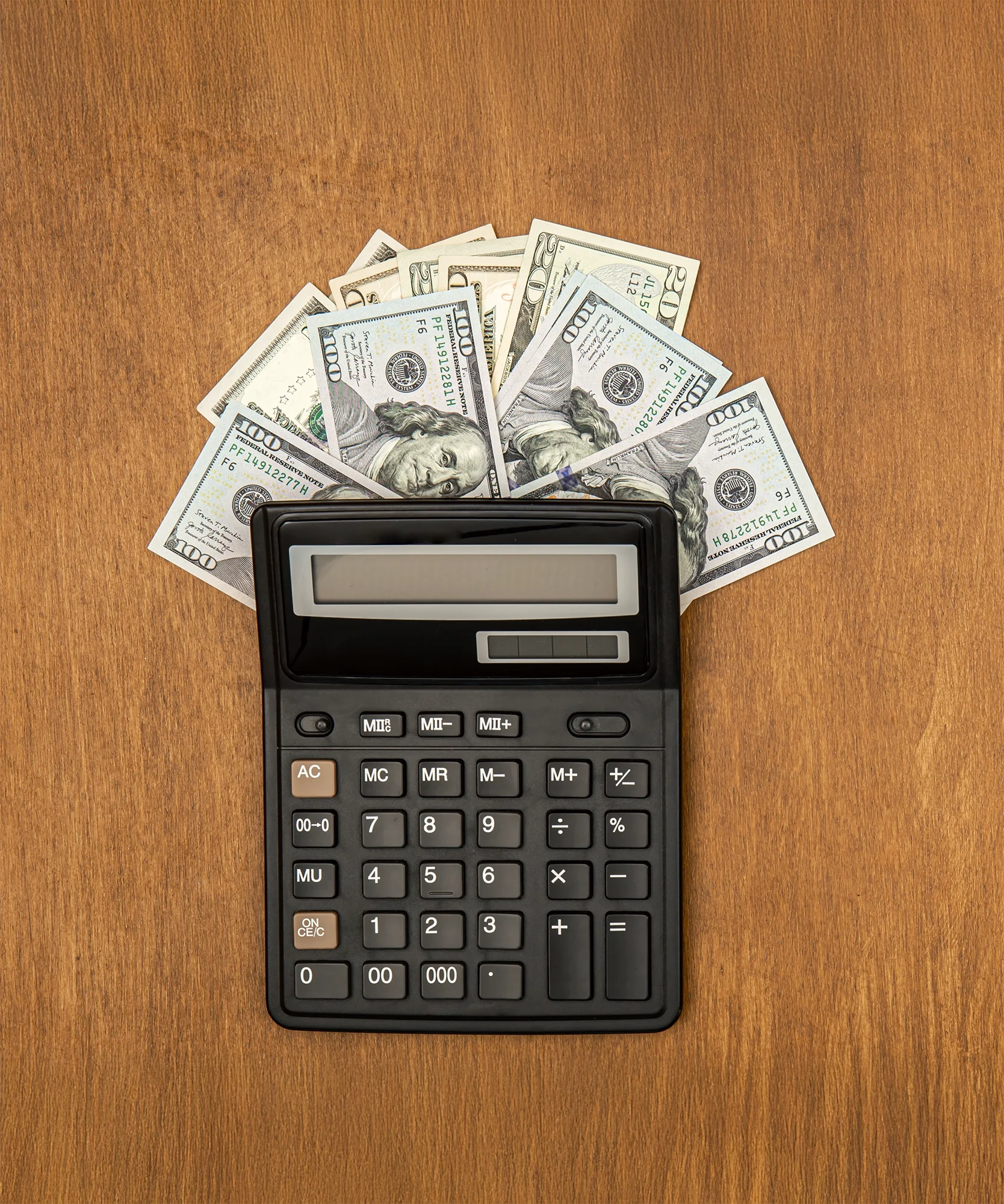 A calculator on a wooden desk with scattered U.S. currency bills including twenties, hundreds, and others around it.