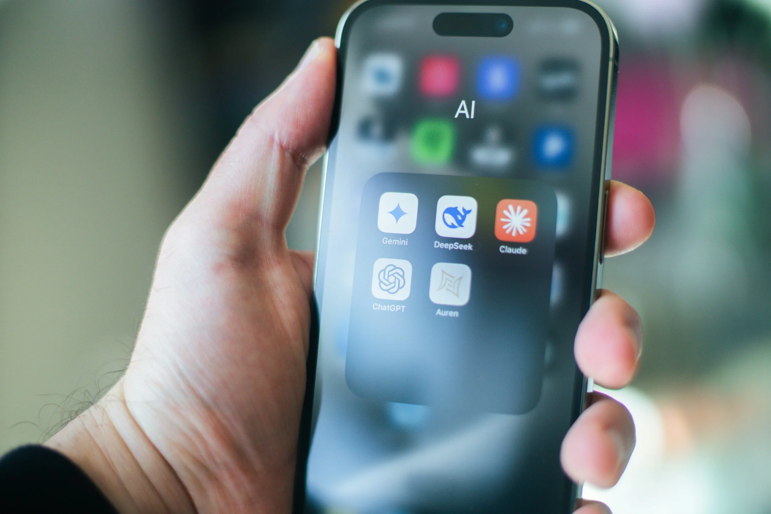 A hand holding a smartphone with a folder labeled 'AI' open on the screen, showing app icons such as Gemini, DeepSeek, Claude, ChatGPT, and Auren.