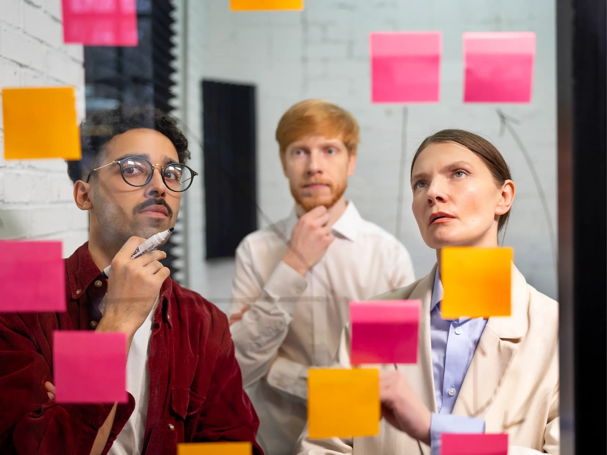 Three professionals studying a glass board covered with colorful sticky notes while discussing ideas and planning strategy, representing collaboration, sales planning, and problem-solving in a business setting.