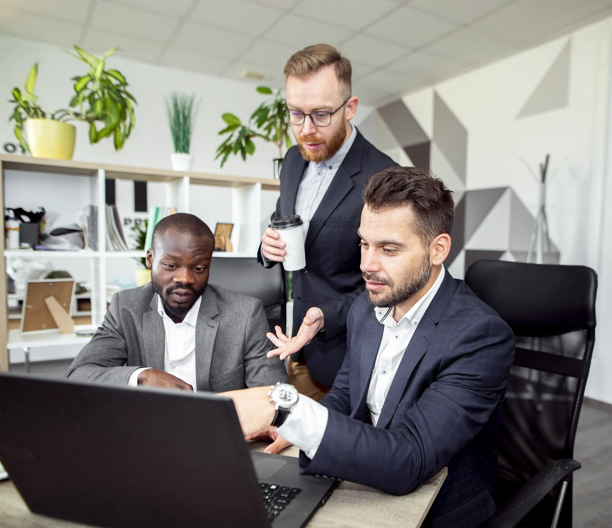 Four business men collaborating in an office, looking at a computer screen and discussing. One man is holding a coffee cup, wearing glasses, and standing, while the others are sitting at the table.