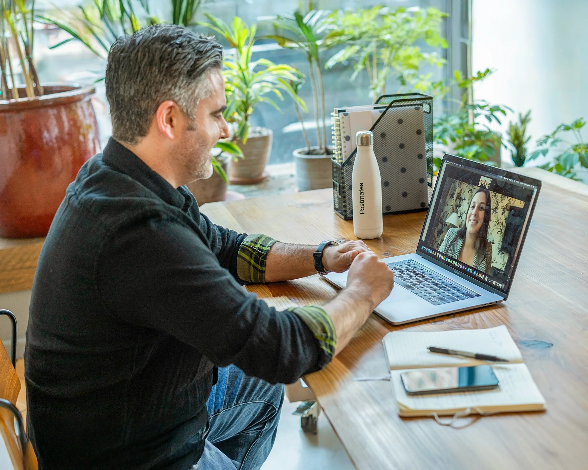 Man sitting at a wooden table during a video call on a laptop, with notebooks, a smartphone, and a water bottle on the table, surrounded by green plants and natural light.