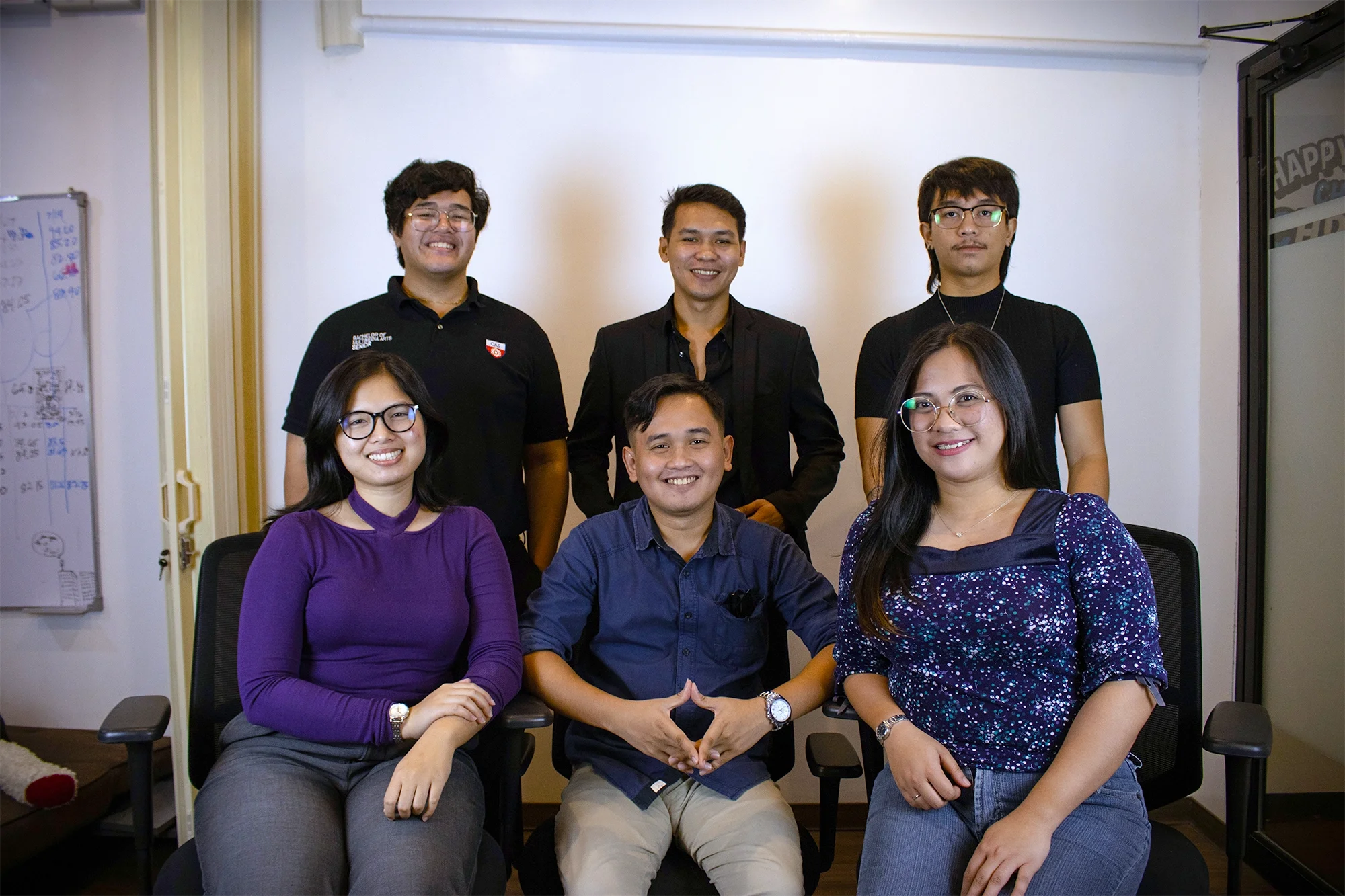 Group of six young adults posing for a photo in an office, sitting and standing in front of a white wall, with a whiteboard on the side.