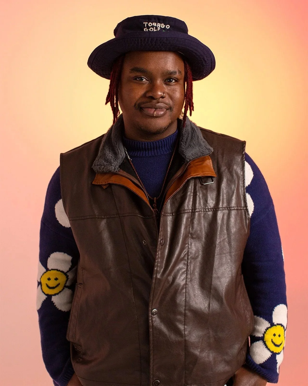 a dark skinned person in a blue brimmed hat, blue jumper and brown vest smiles into the camera