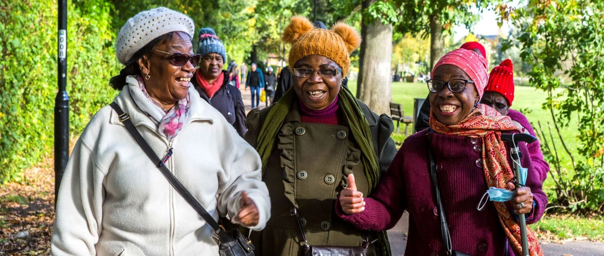 Group of four elderly women, smiling and walking in a park, enjoying fall weather, wearing colorful hats, scarves, and jackets.