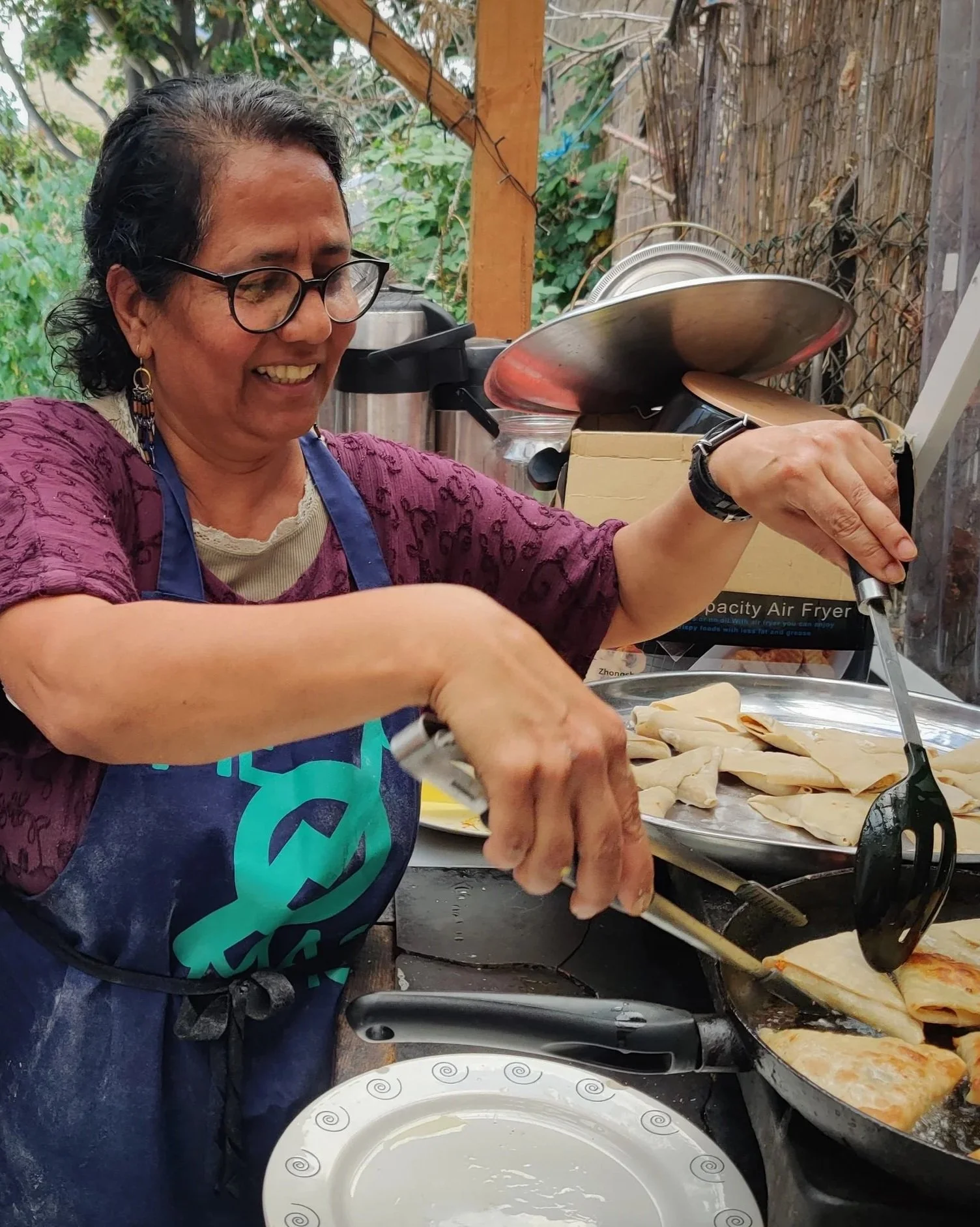 a woman in a purple shirt and blue apron cooks in an outdoor kitchen