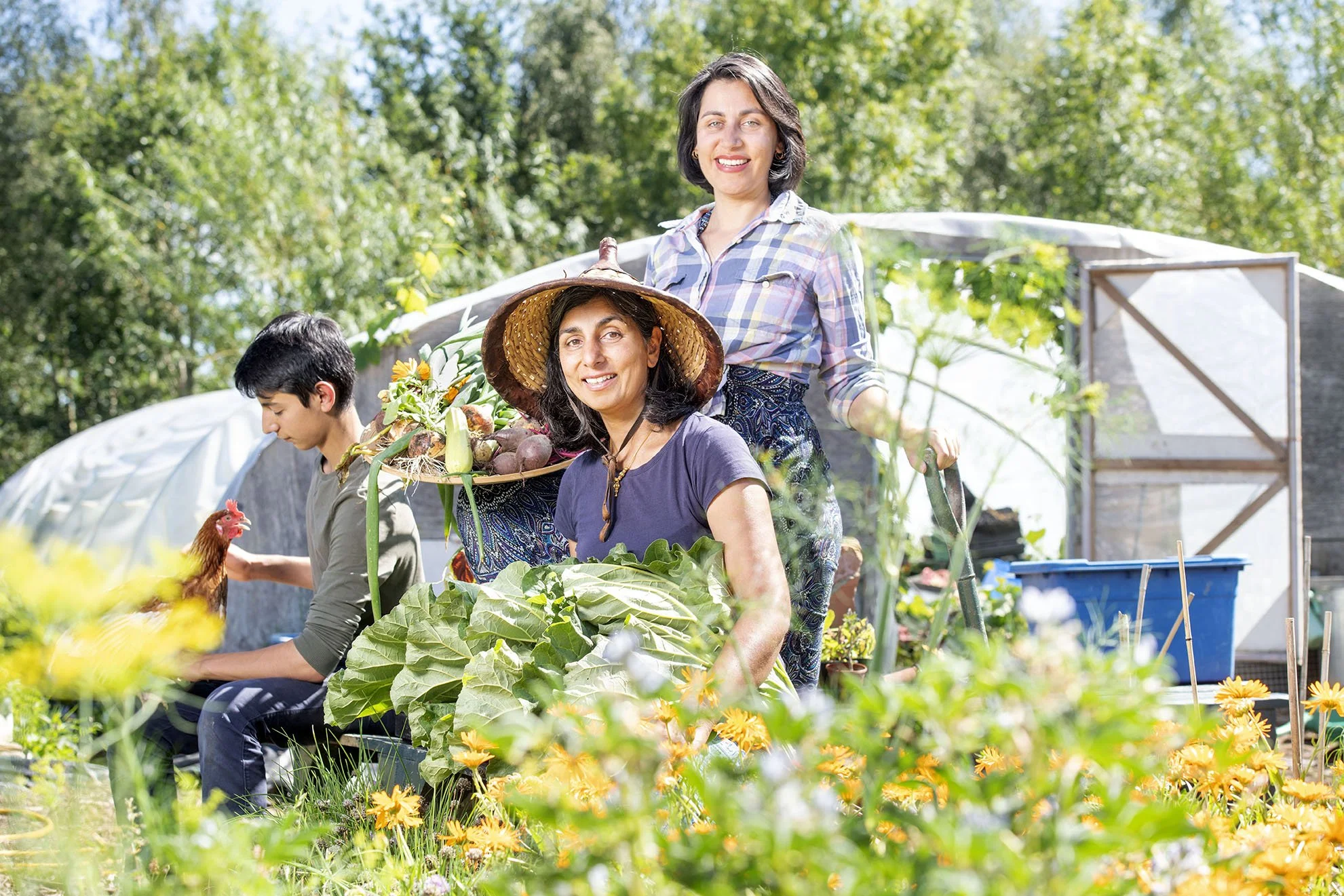 two women smile at the camera in front of a polytunnel while a young man next to them looks at a chicken