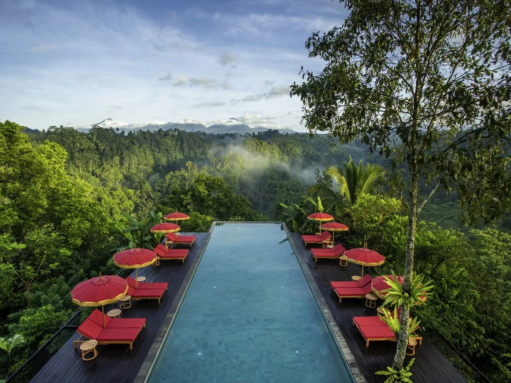 Infinity pool with red umbrellas and loungers overlooking lush green forested hills.