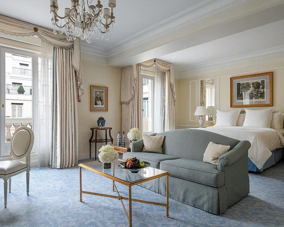 Luxury hotel suite with elegant decor, featuring a bed with white linens, a blue sofa, a glass coffee table with fruit and flowers, a chandelier, framed artwork, and large windows with drapes.