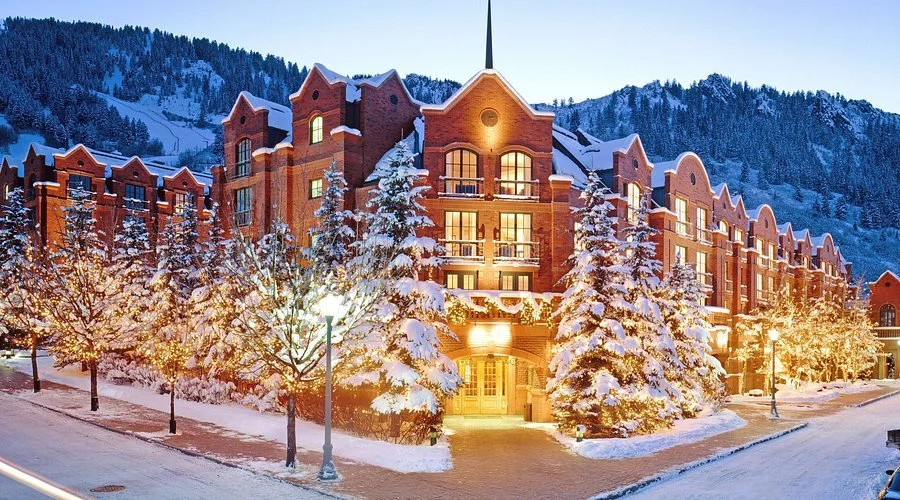 A snow-covered luxury hotel building with a red brick facade, surrounded by snow-laden trees and mountains in the background. The entrance is warmly lit and the scene conveys a wintery, picturesque setting.