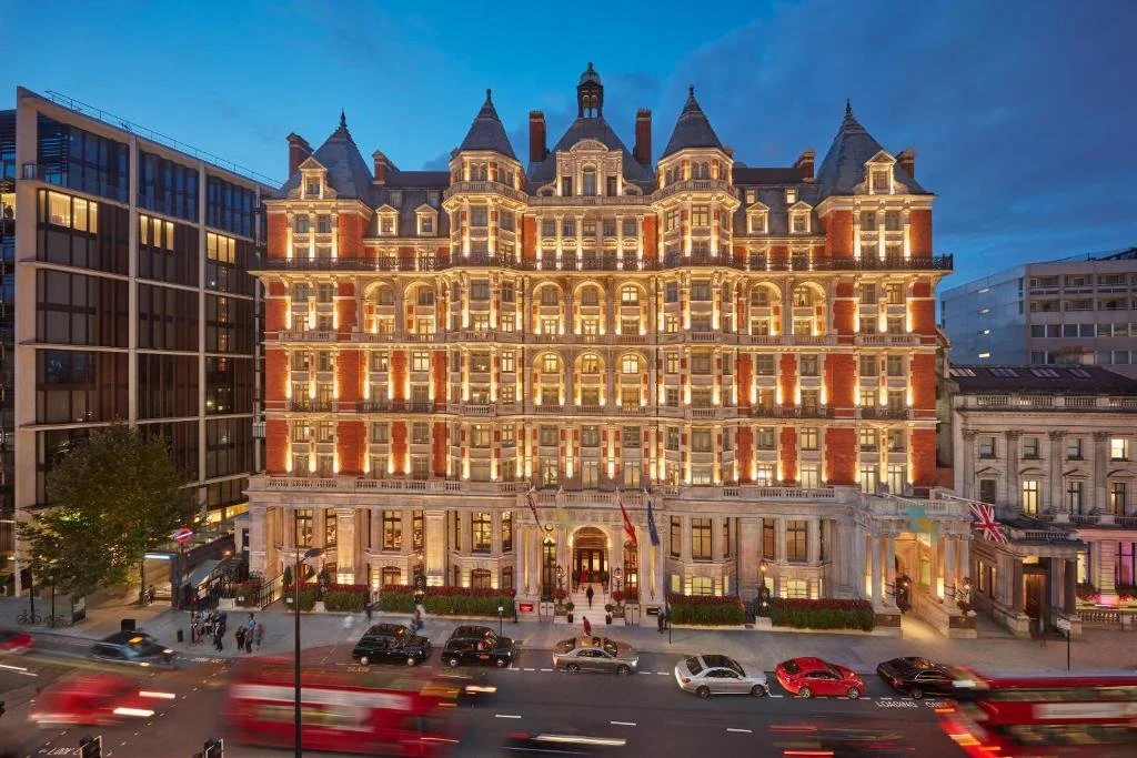 Illuminated hotel building at night with a Victorian architectural style, featuring multiple gabled rooftops. The street in front has moving red double-decker buses and several cars.