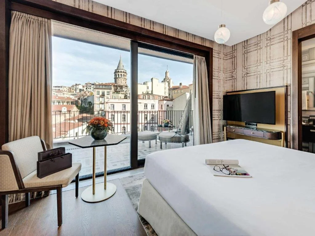 Hotel room interior with large bed, TV, chair, and table; balcony view of cityscape and historic tower in distance.