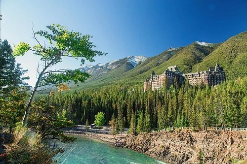 Banff Springs Hotel surrounded by forest with mountains in the background and a river in the foreground on a sunny day.