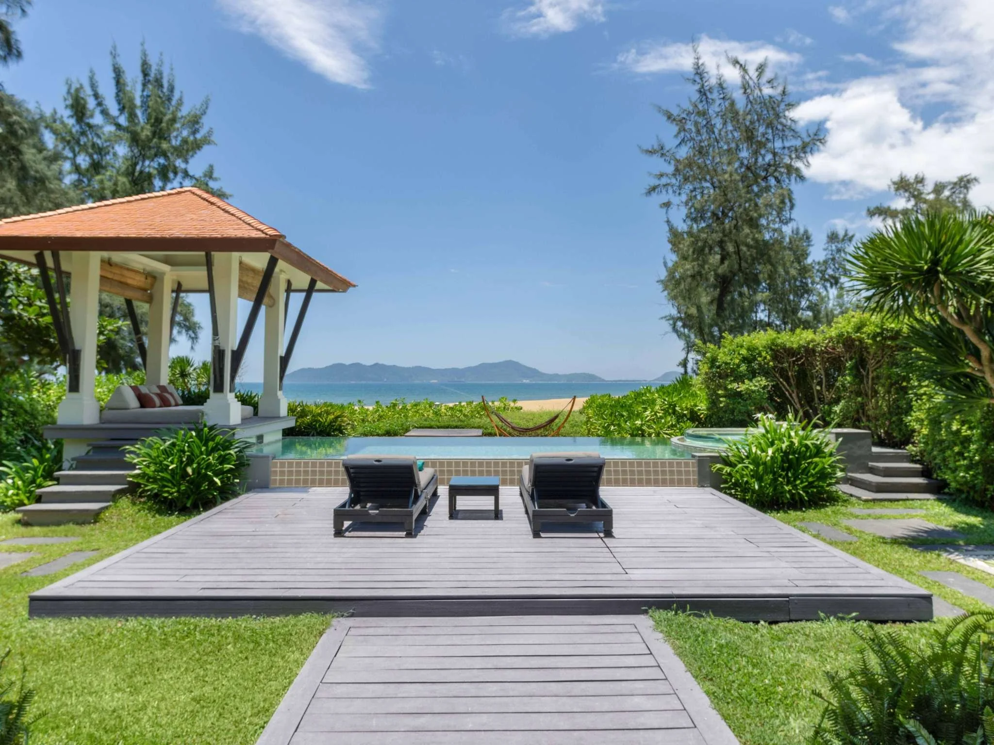 Luxury outdoor lounge area with a pool, lounge chairs, and gazebo overlooking a beachfront with ocean view and mountains in the background.