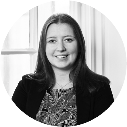 Black and white portrait of a young woman with shoulder-length hair, smiling, wearing a patterned top and a blazer, seated in front of a window with curtains.