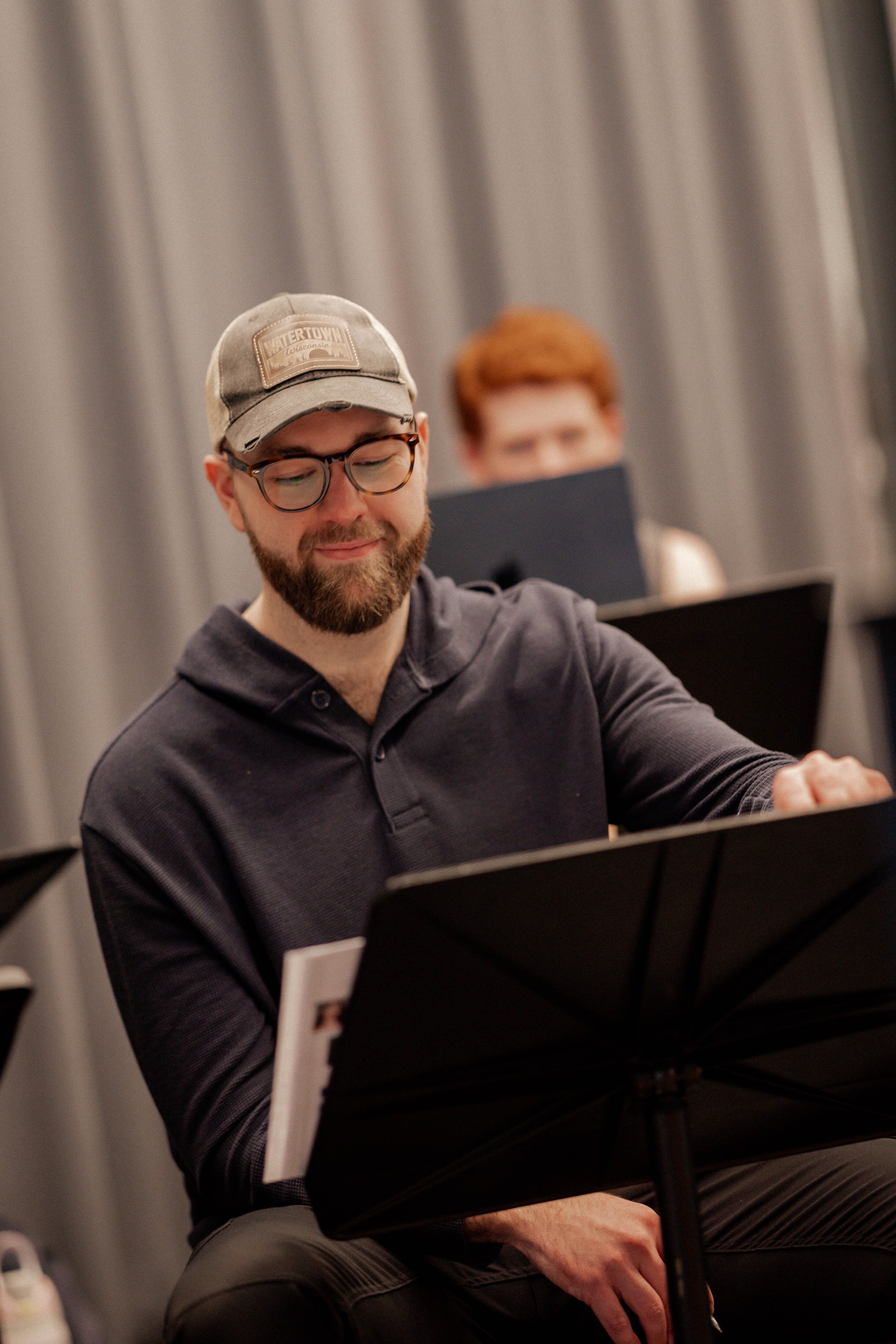 A man with glasses, a beard, and a baseball cap sitting in front of a music stand, smiling softly, with a woman with short red hair and glasses working on a laptop in the background.
