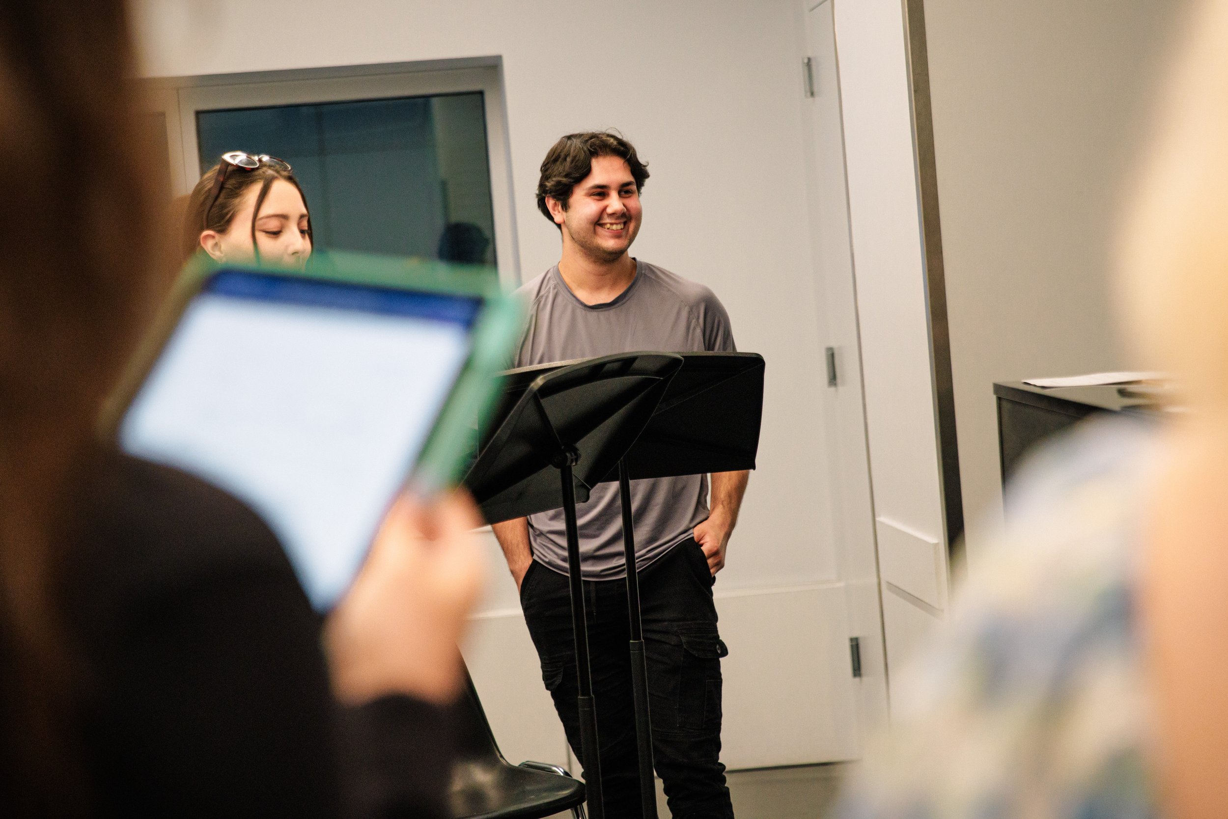 A young man with dark hair, wearing a gray T-shirt, is smiling and standing behind an empty music stand. Several people are in the foreground, one is holding a phone, and another has glasses on her head. The setting appears to be an indoor room, poss