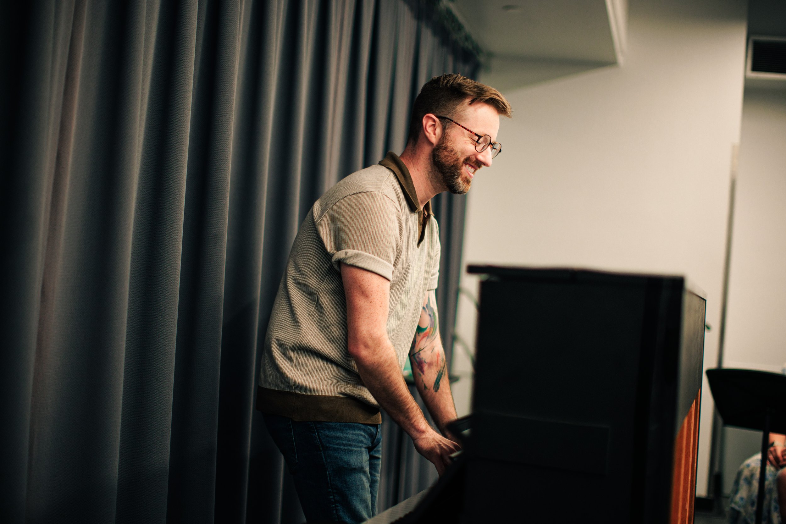 A man with glasses and a beard, smiling while playing a piano in a room with gray curtains.