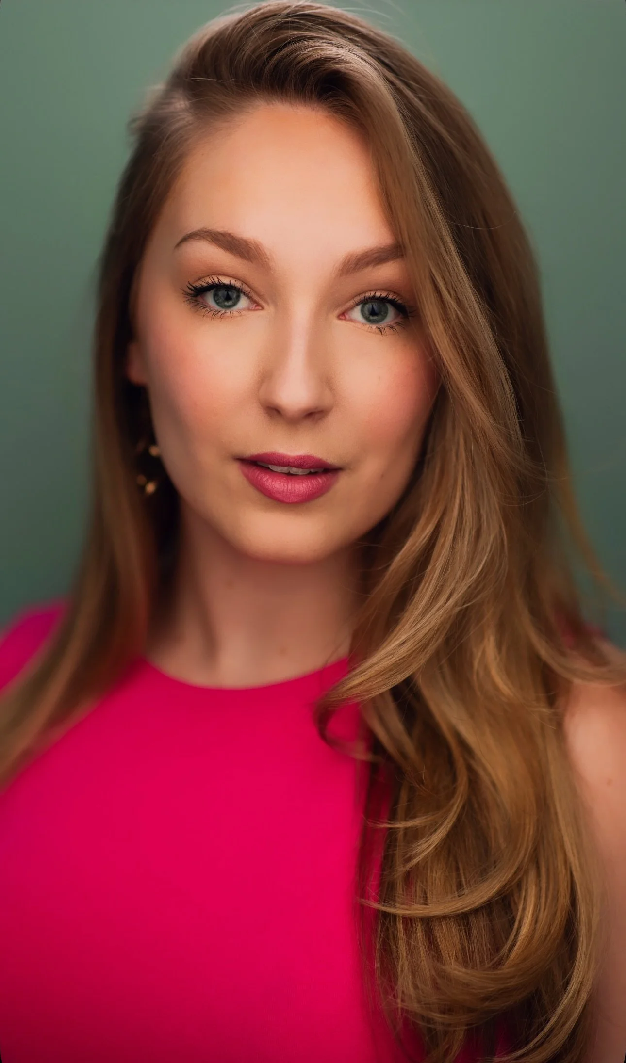 Close-up portrait of a young woman with long, wavy, light brown hair, blue eyes, wearing a bright pink top, against a green background.