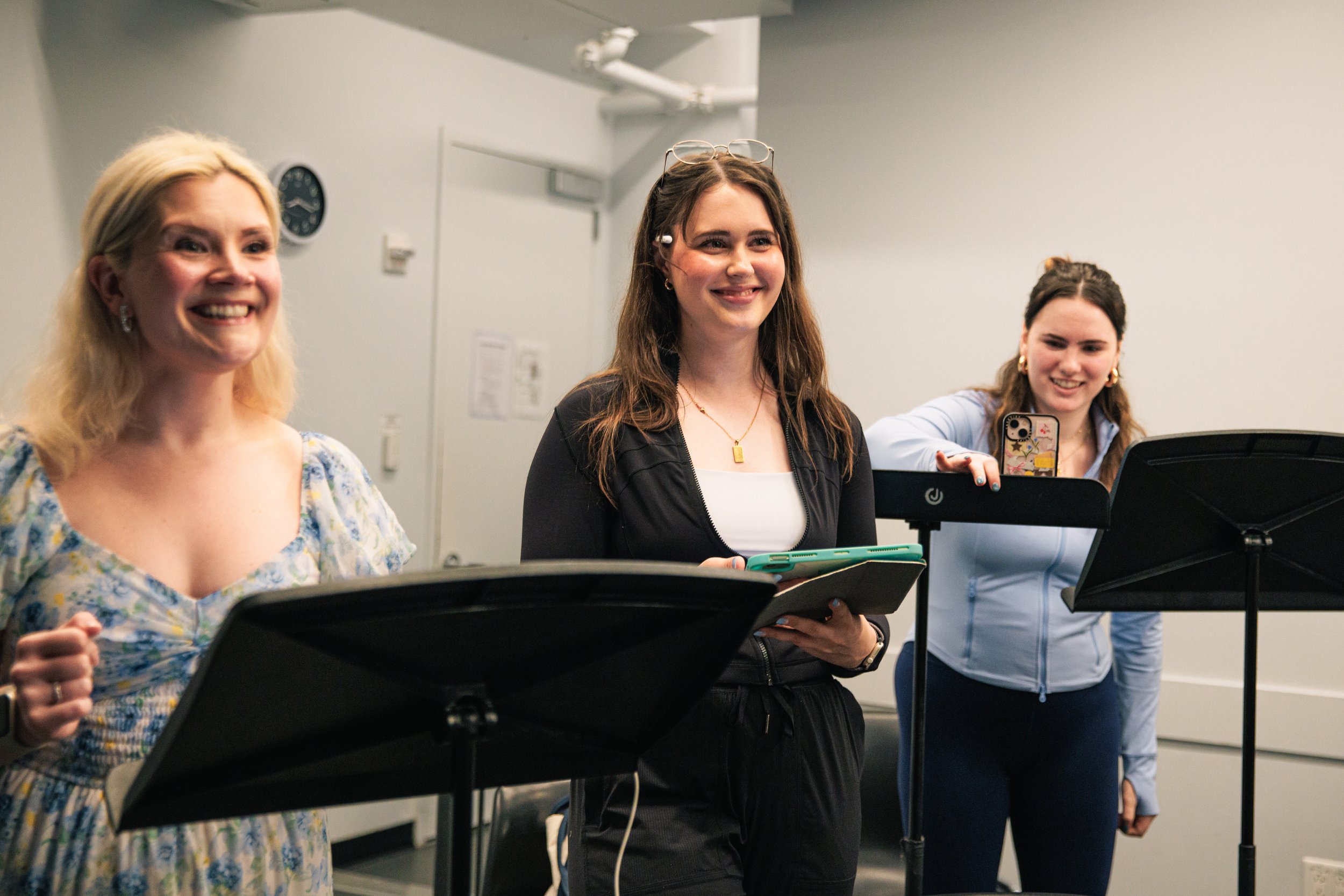 Three women standing in front of music stands, smiling and posing for a photo in a room with white walls.