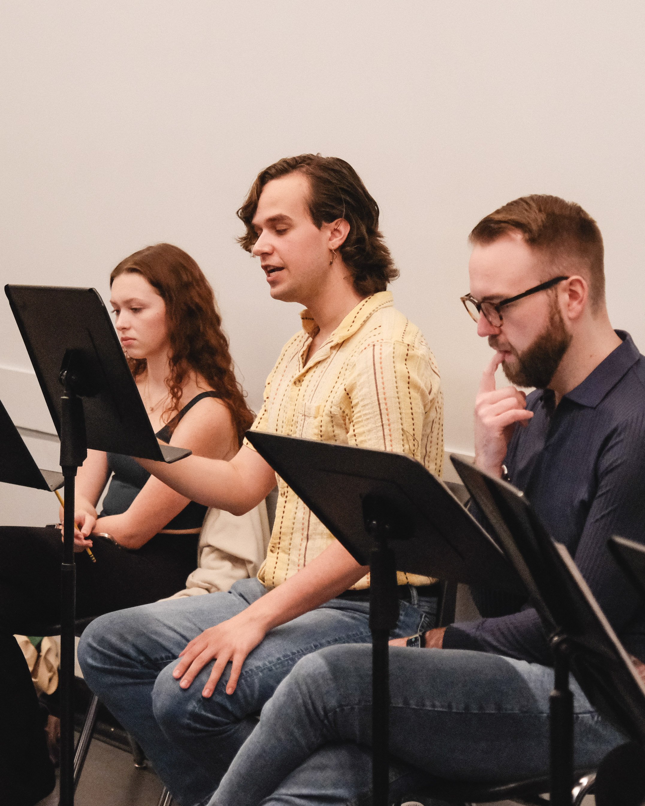 Three people sitting in a row, reading sheet music on black music stands, during a music rehearsal or class.