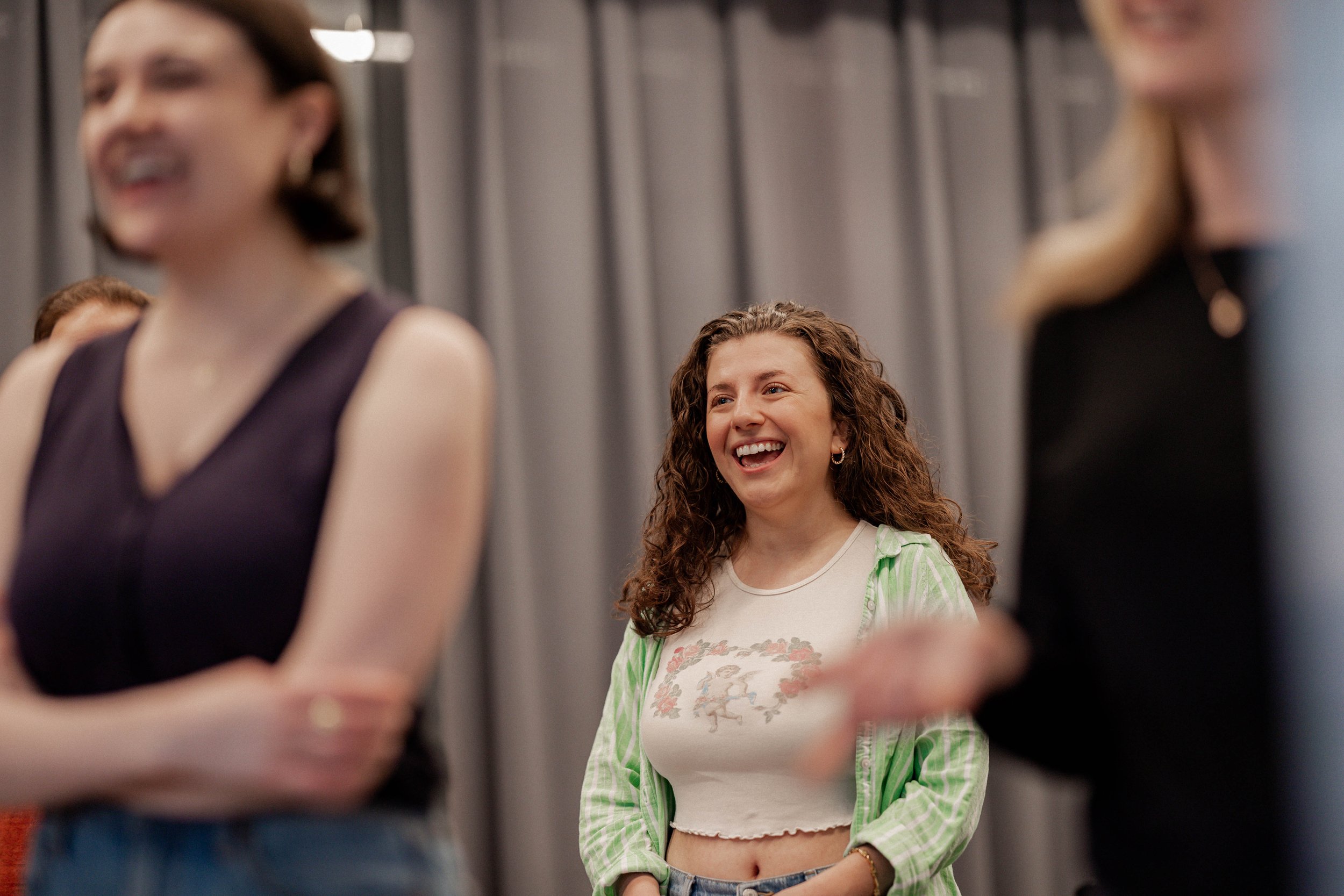 A woman with curly hair and a green striped shirt smiling and laughing among a group of women at an indoor gathering.