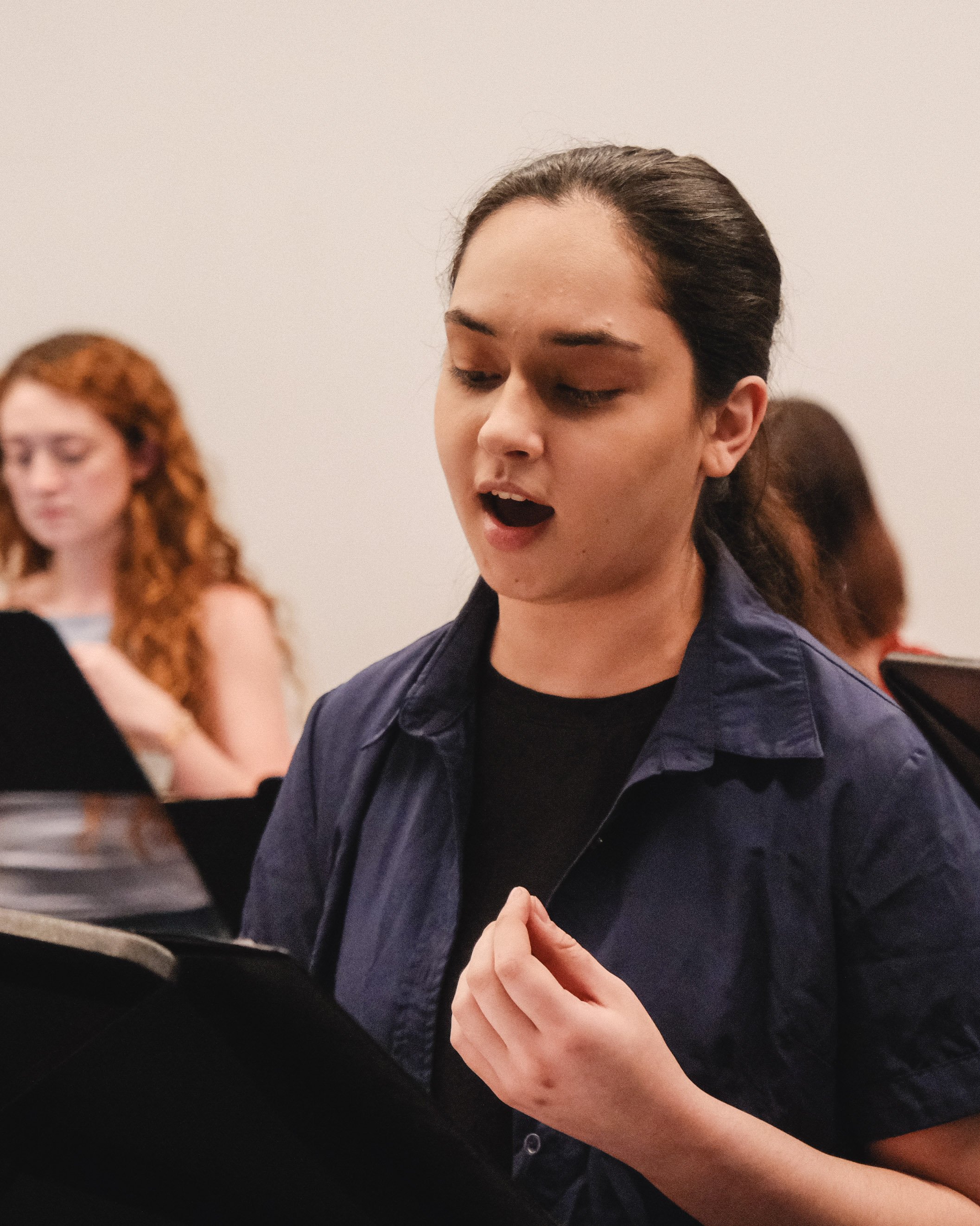 A young woman with dark hair singing or speaking in a choir or rehearsal, with other women in the background.