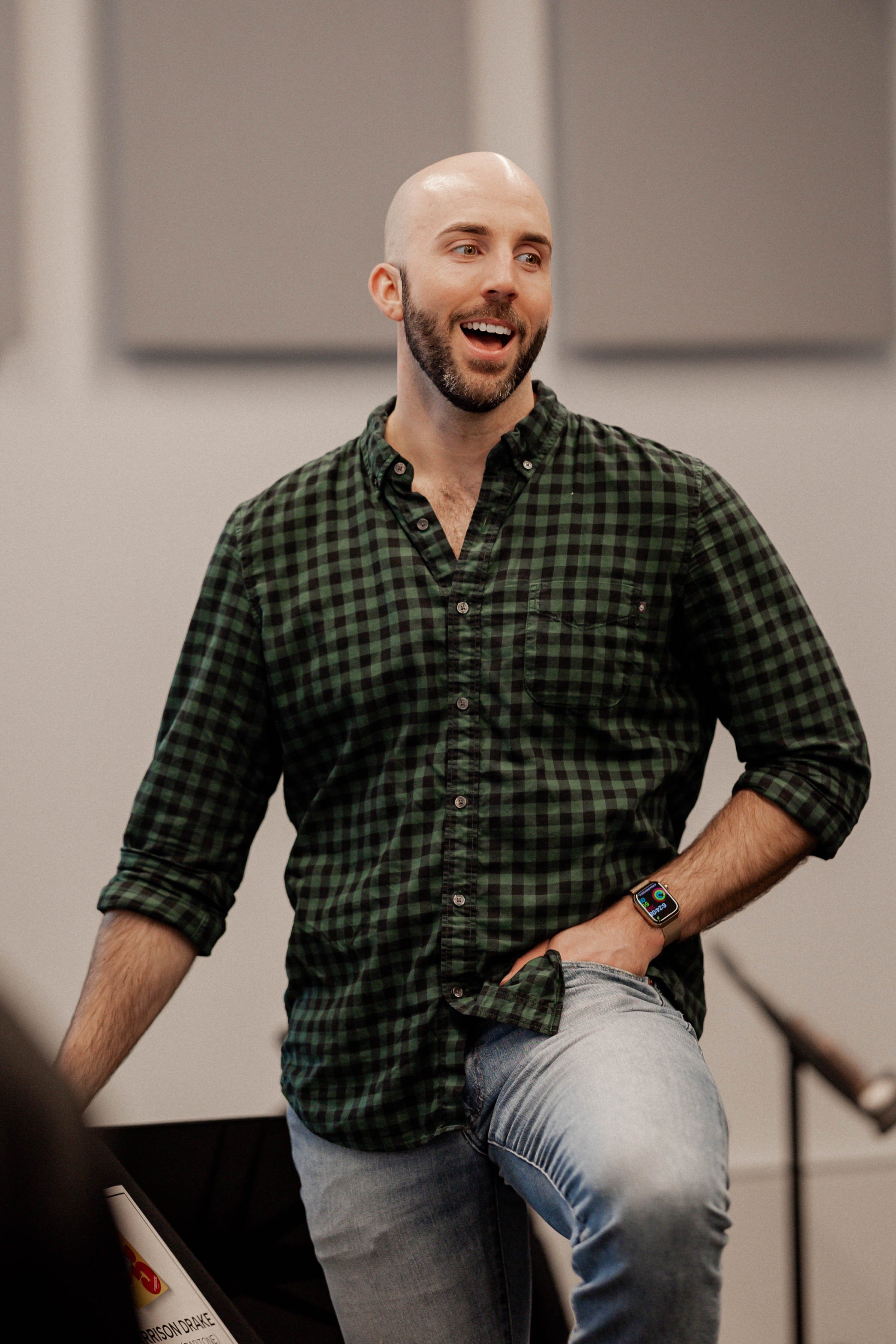 A man with a beard and shaved head smiling, wearing a green and black checkered shirt and light blue jeans, with a smartwatch on his left wrist, standing in a room that appears to be a music or choir rehearsal space.