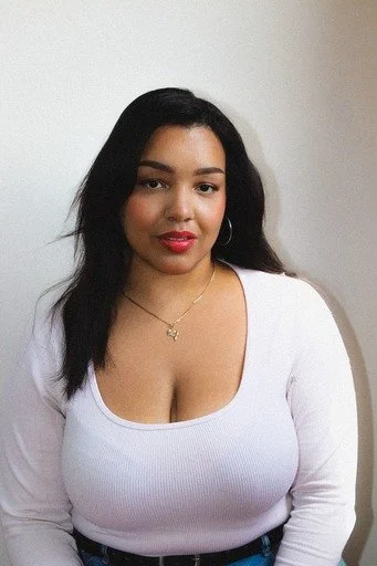 Portrait of a young woman with dark hair, wearing a white top and gold jewelry, standing against a plain wall.