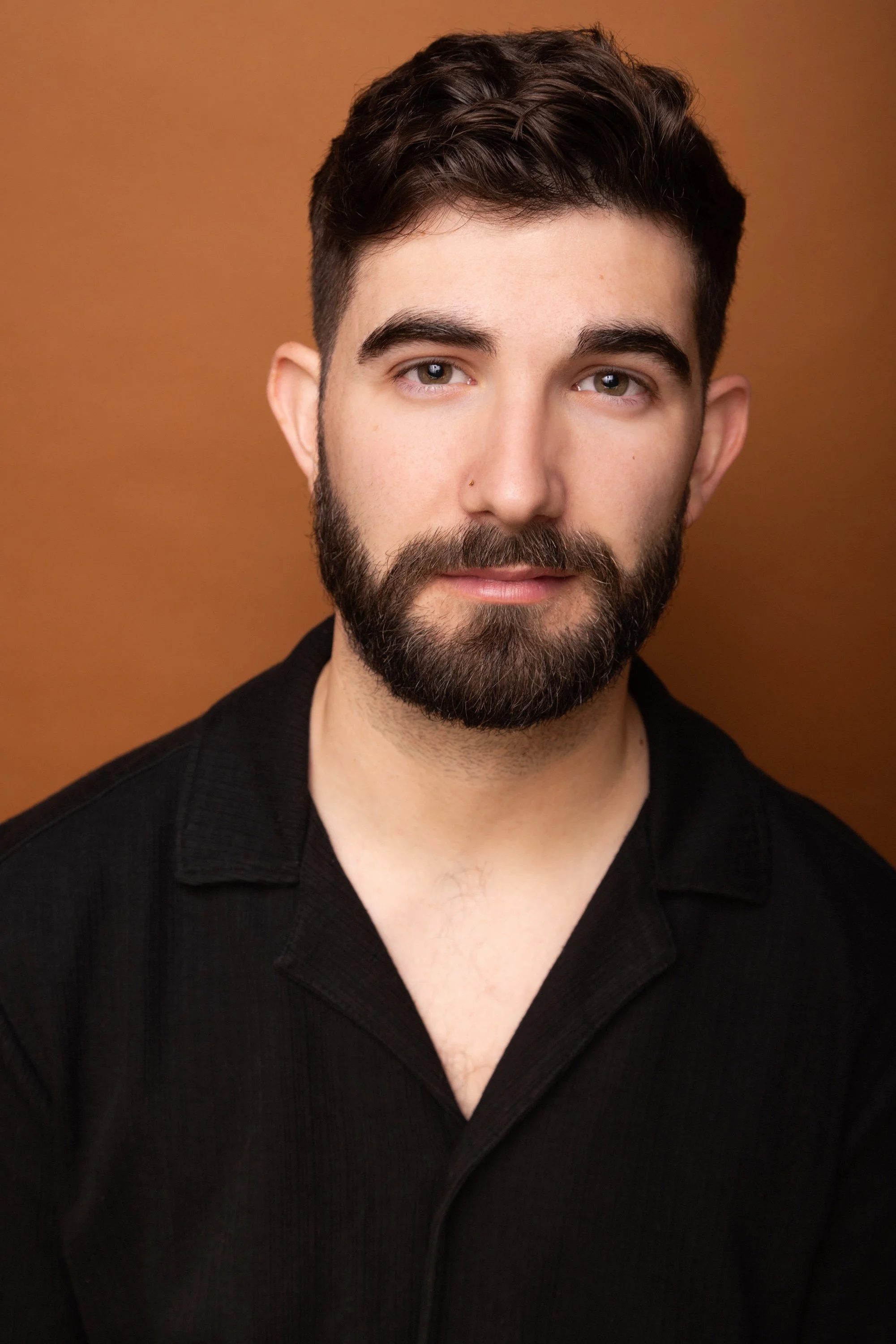 Portrait of a young man with dark hair, a beard, and hazel eyes, wearing a black shirt, against a brown background.