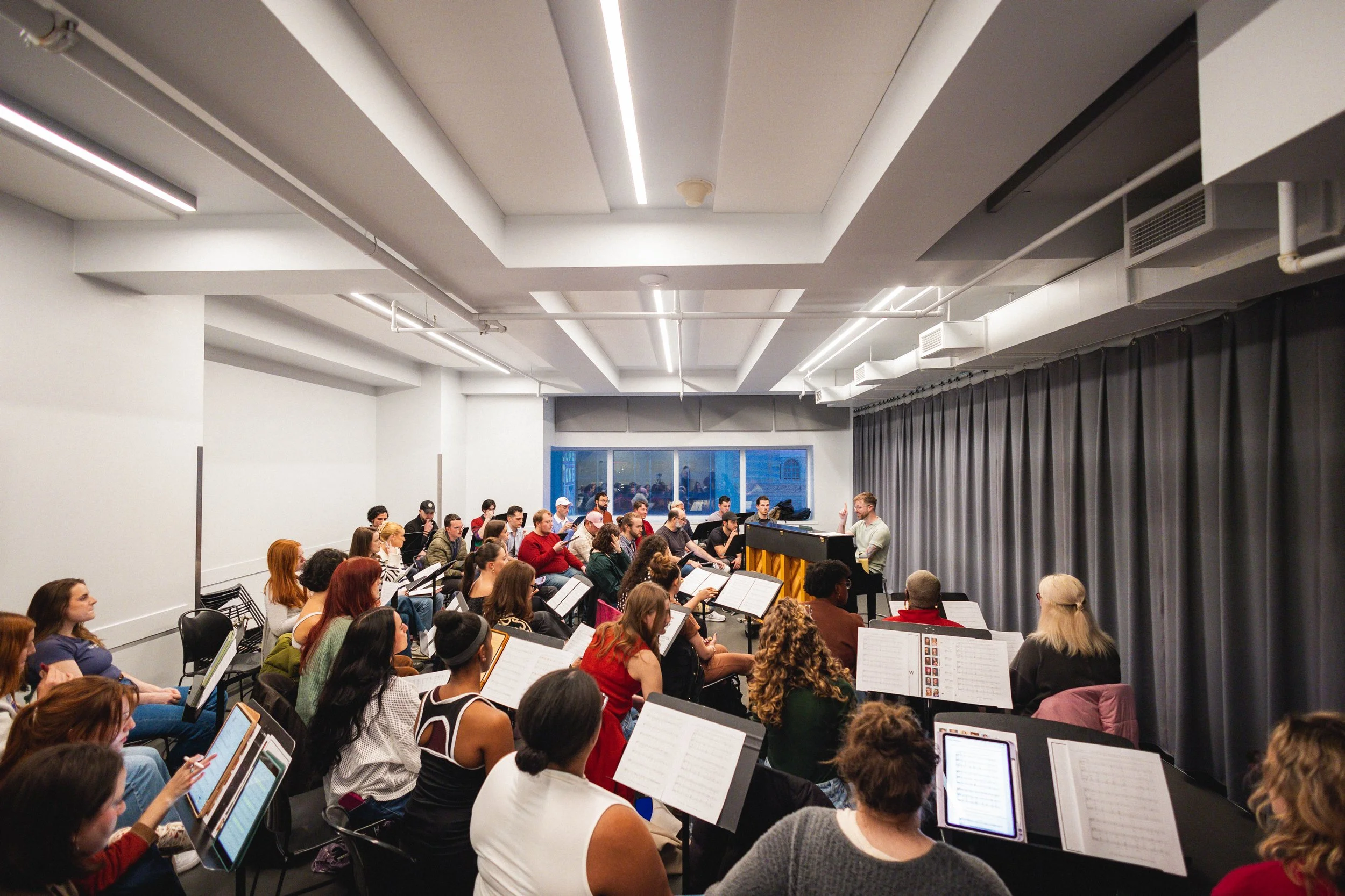A large group of people in a music rehearsal room, seated with music sheets, facing a conductor who is standing and conducting at a piano. The room has white walls, grey curtains, and ceiling lights.