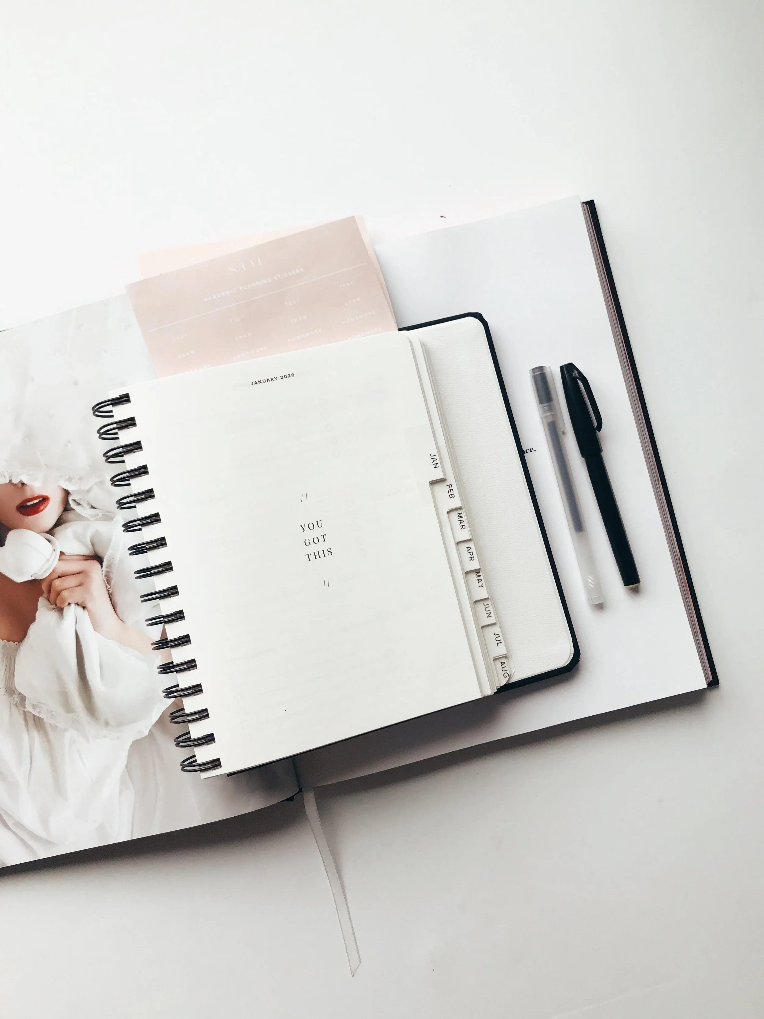 Un agenda ouvert avec des pages blanches, un stylo noir, un carnet à onglets avec les mois, une photo d'une femme avec des lèvres rouges et un ruban en papier