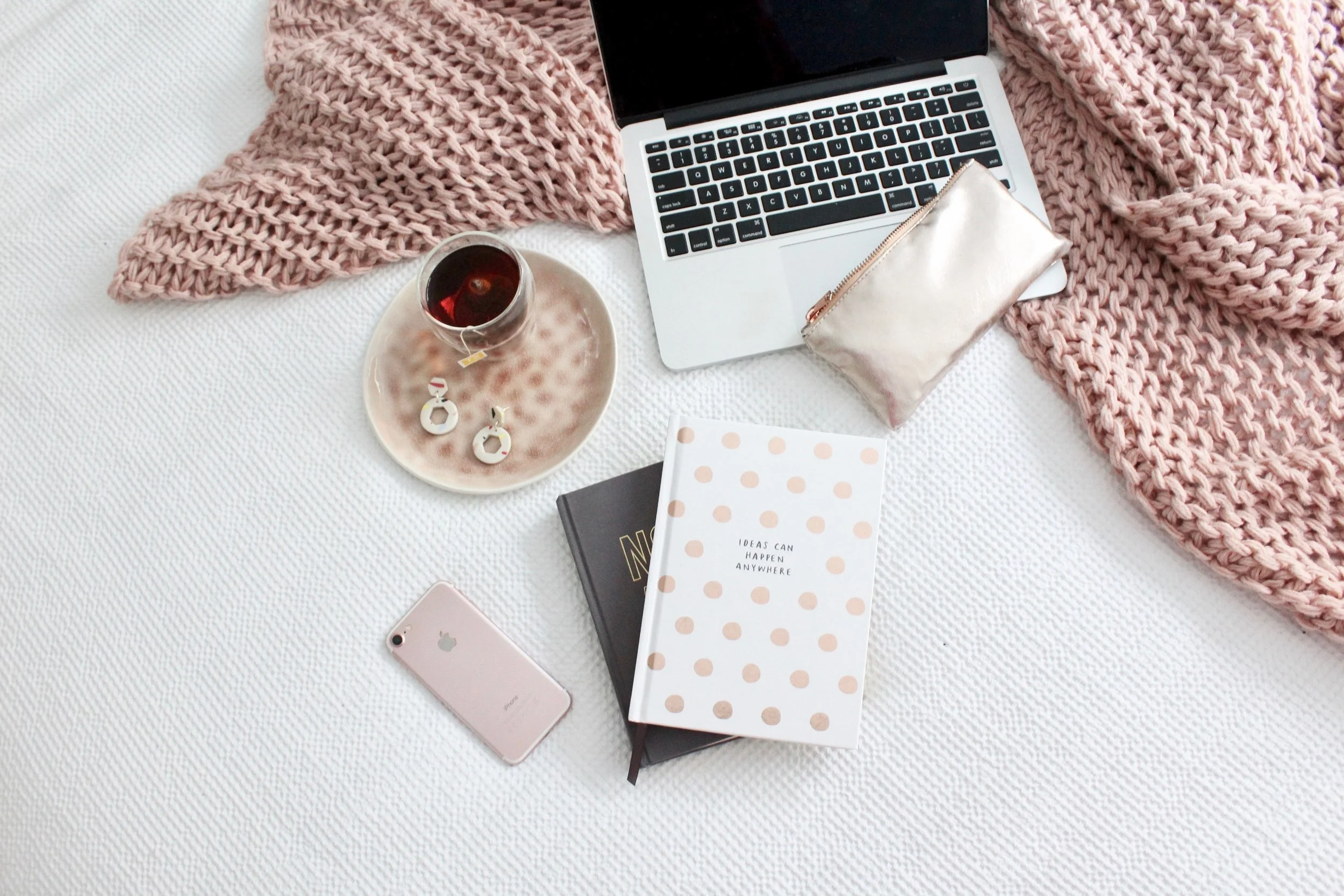 Surface blanche avec un ordinateur portable, un mug avec une boisson chaude, une paire de boucles d'oreilles en forme de cercle, un carnet à pois avec la phrase "Ideas can happen anywhere", un iPhone rose, un sac en satin, une tasse en porcelaine avec une boisson chaude, une écharpe en tricot rose.