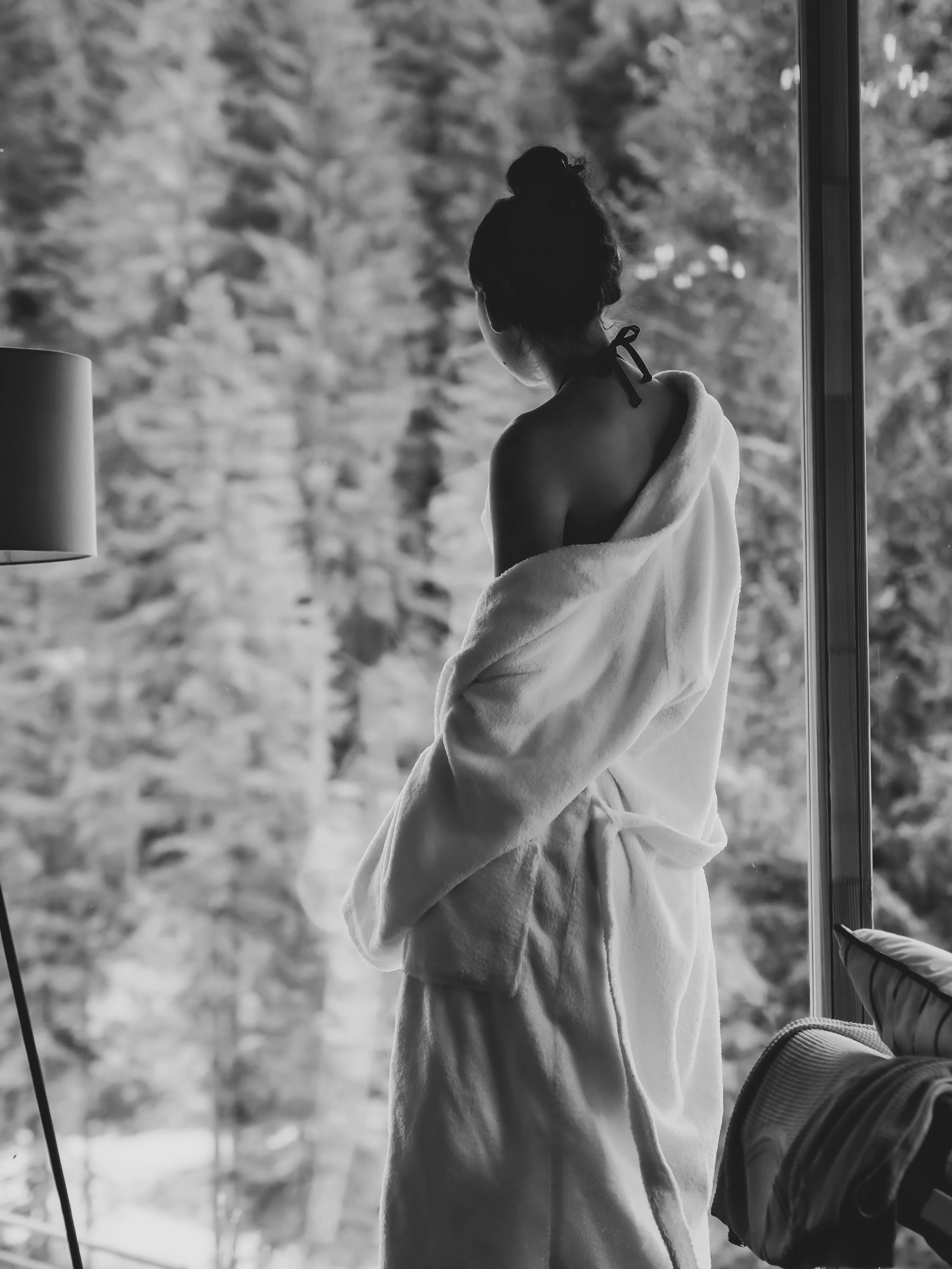 Une femme vue de dos, vêtue d'une robe en peignoir, regardant par une fenêtre, avec des arbres en arrière-plan.
