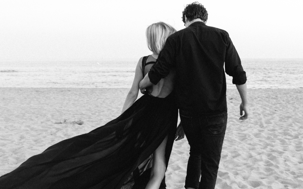 Un couple marche sur une plage, vue de derrière, en noir et blanc.